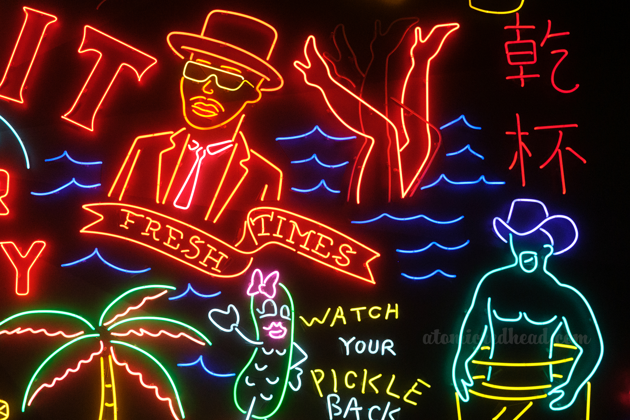 A portion of the massive neon mural at Grand Central Market which features a man in a suit, top hat, and glasses, a pair of legs in water, a pickle with a face that reads "Watch Your Pickle Back" next to it, and a cowboy.