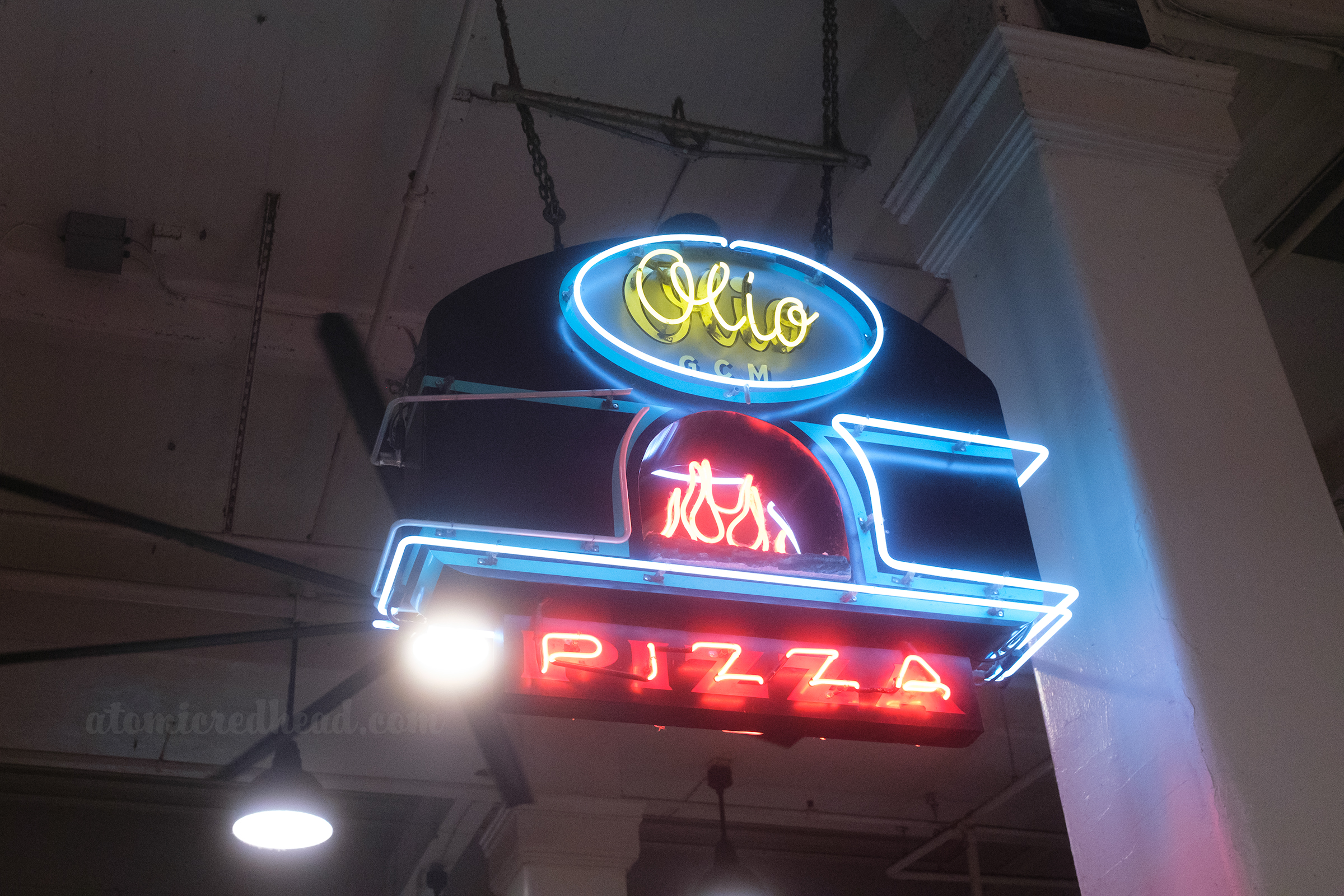 Sign for Olio pizza, which reads "Olio" in yellow neon scrip" neon flames, and "Pizza" in red letters.