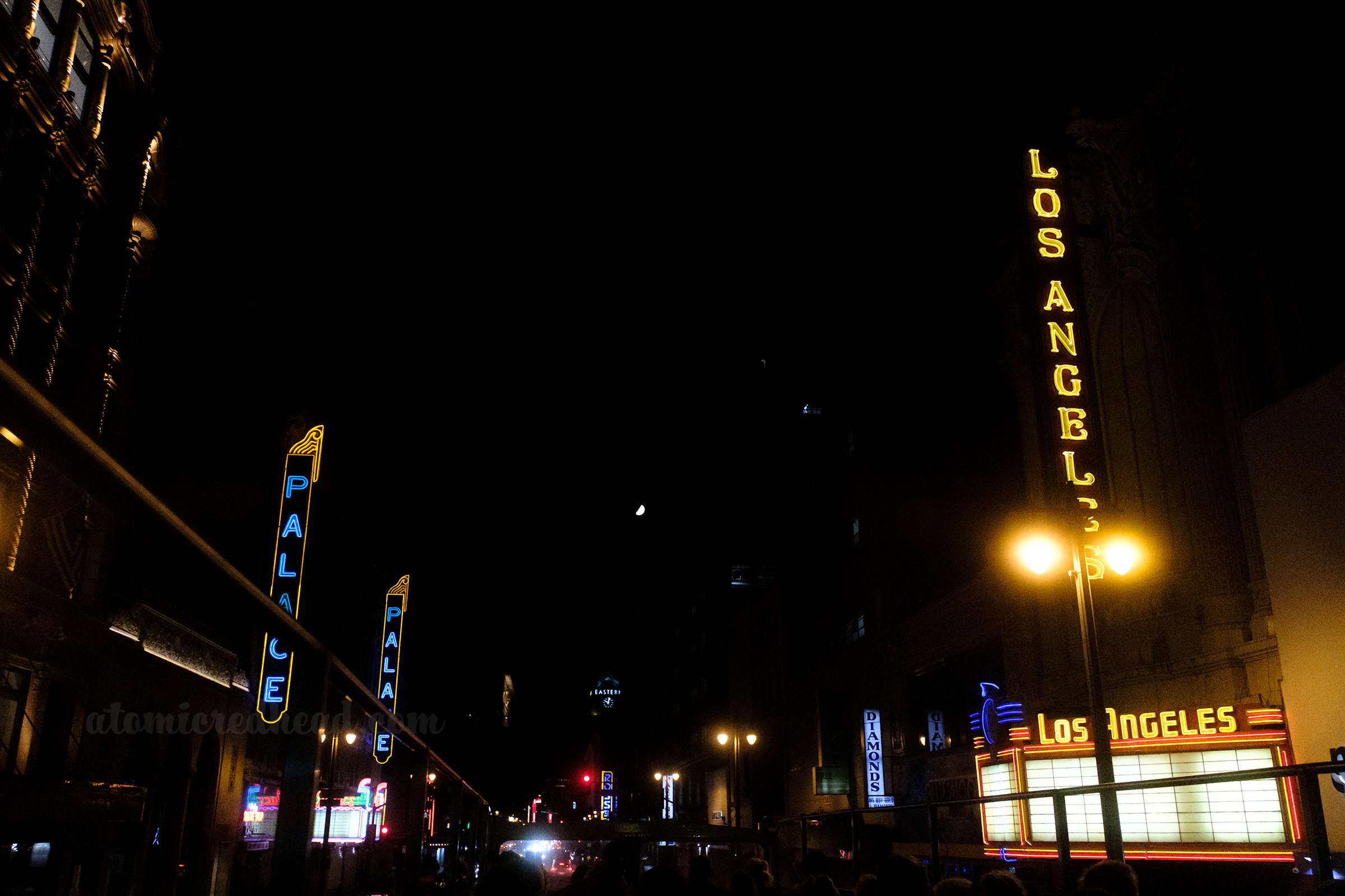 View down Broadway, with a variety of blade style neon signs for both active and defunct theaters. One reads "Los Angeles" in yellow neon, another reads "Palace" in blue. Further down a sign for a Ross Dress for Less is done in the same style to fit in.