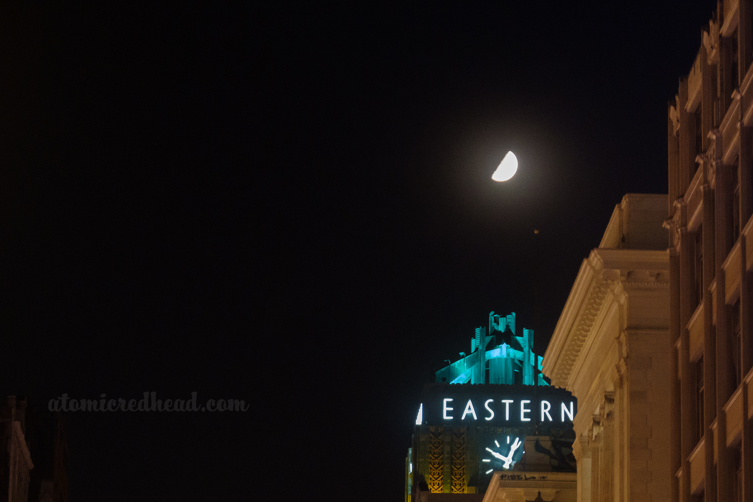 Top of the Art Deco Eastern building which reads "Eastern" in neon, and a neon clock below, the half moon sits above in the sky.