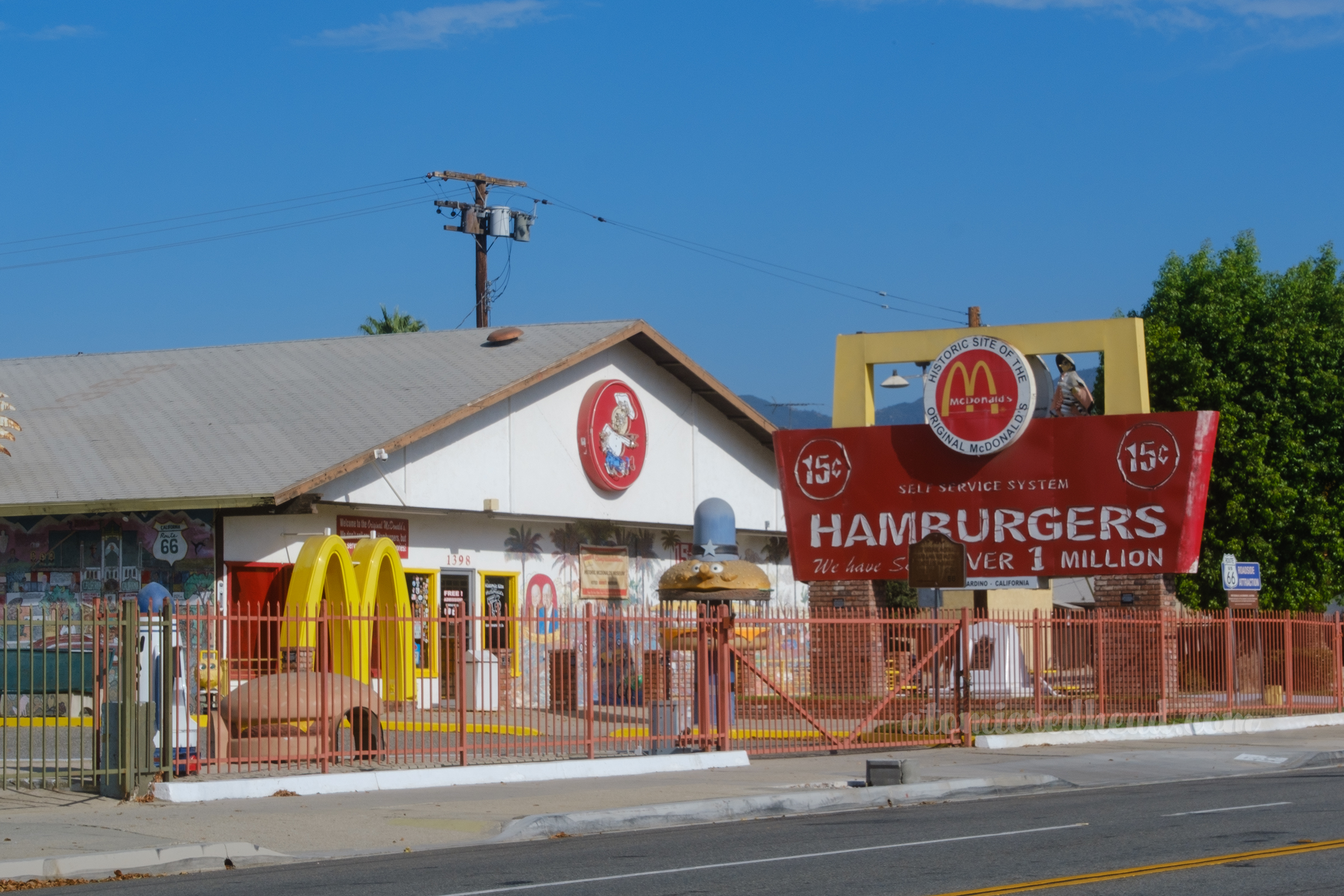 Overall view of the McDonald's Museum, which features a large red and yellow sign reading "Historic Site of the Original McDonald's Self-Service System 15 Cent Hamburgers We have sold over 1 Million." Outside various pieces of playplace equipment stand.
