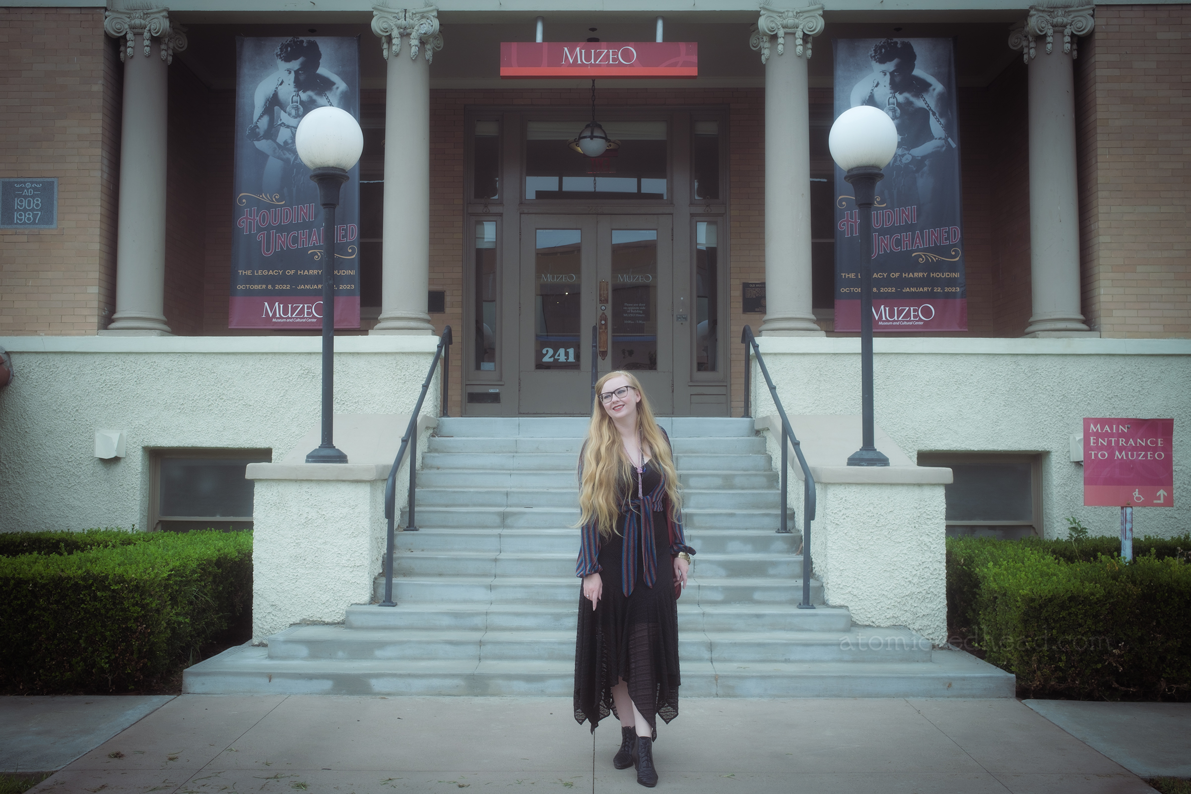 Myself, wearing a multi-colored bolero over a black lace dress, a pink lariat necklace, and a blue glass beaded necklace, and a pair of gold, shackle like bracelets, standing in front of the Muzeo's white brick building.