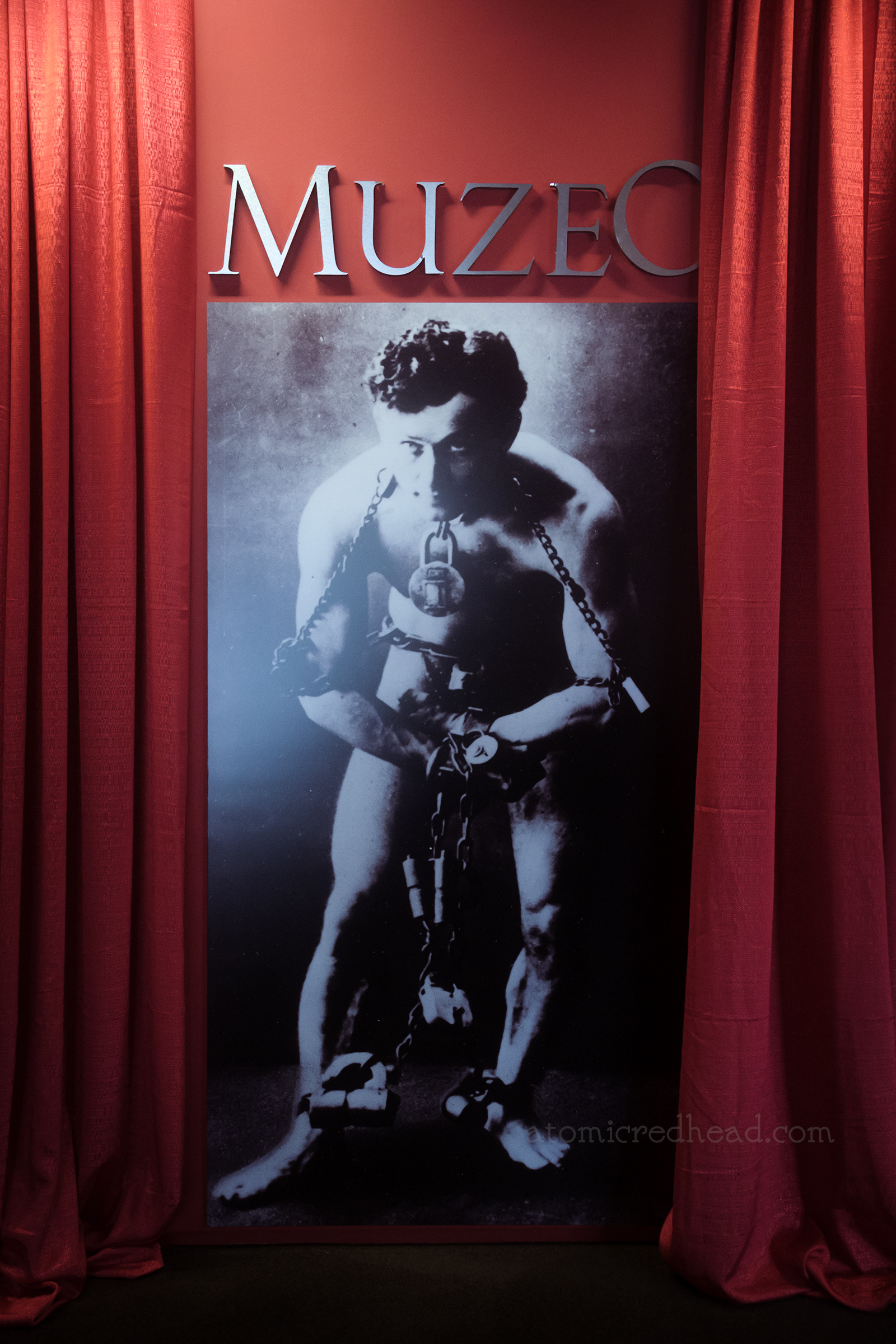 Red curtains flank a black and white photograph of Houdini in shackles, above gold letters read "Muzeo"