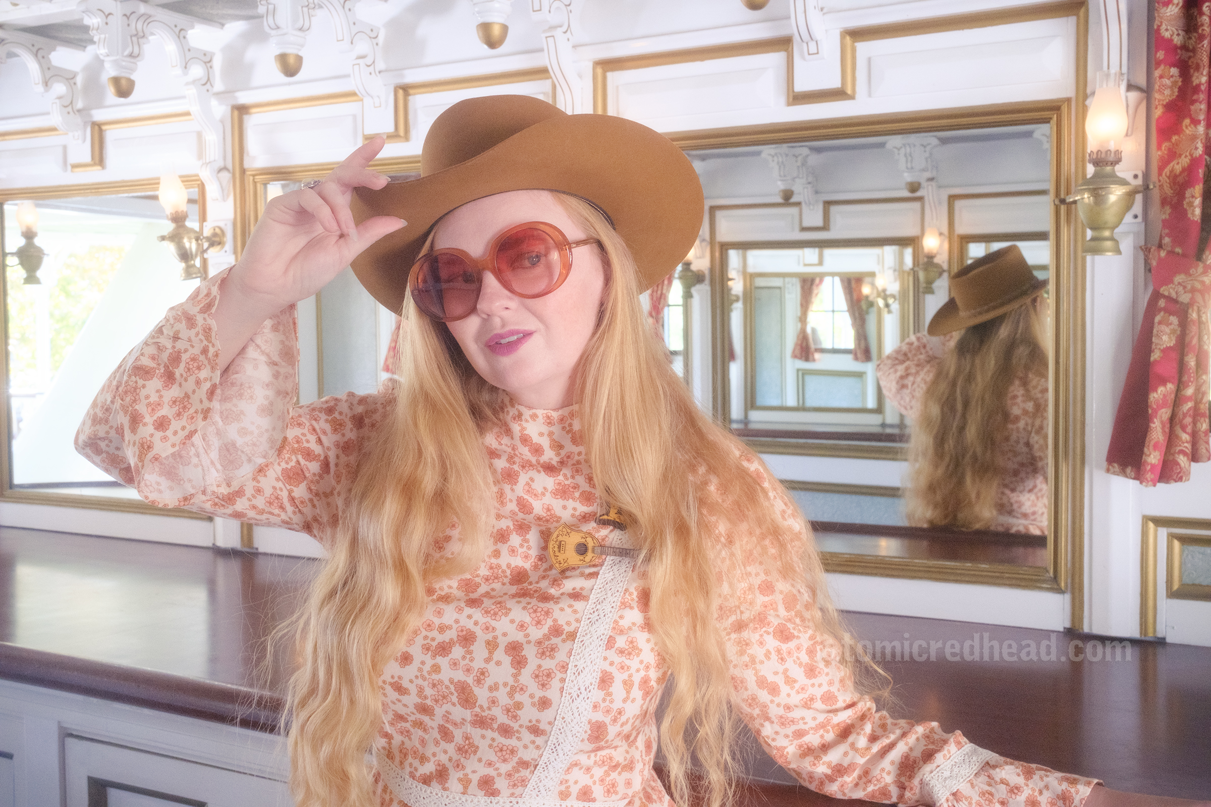 Myself, wearing a light brown cowboy hat, a long sleeve mini dress featuring a floral pattern in shades of orange and brown, with lace detail on the cuffs and waist, and white fringe cowboy boots, leaning against the mirrored bar aboard the Mark Twain Riverboat.