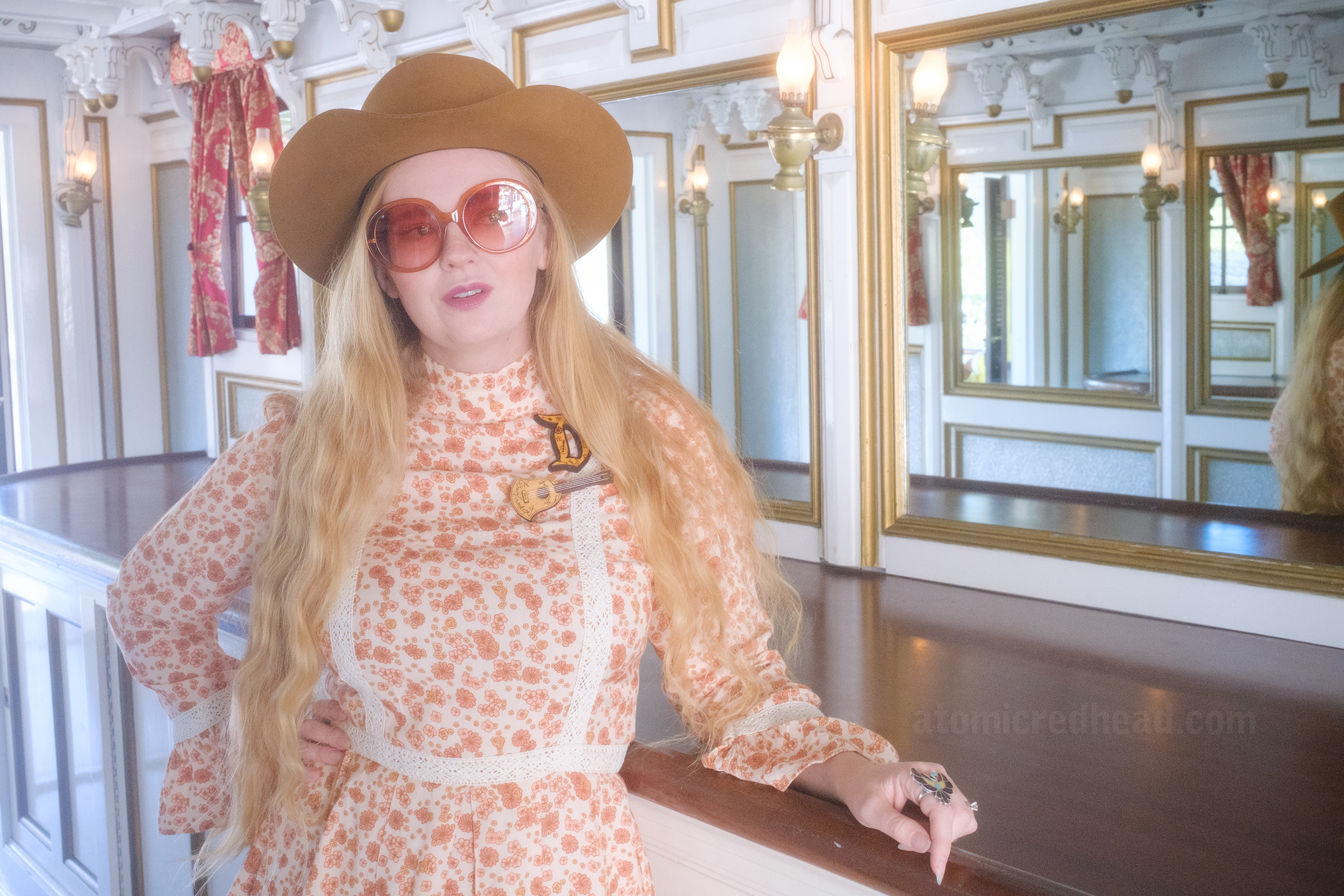 Myself, wearing a light brown cowboy hat, a long sleeve mini dress featuring a floral pattern in shades of orange and brown, with lace detail on the cuffs and waist, and white fringe cowboy boots, leaning against the mirrored bar aboard the Mark Twain Riverboat.