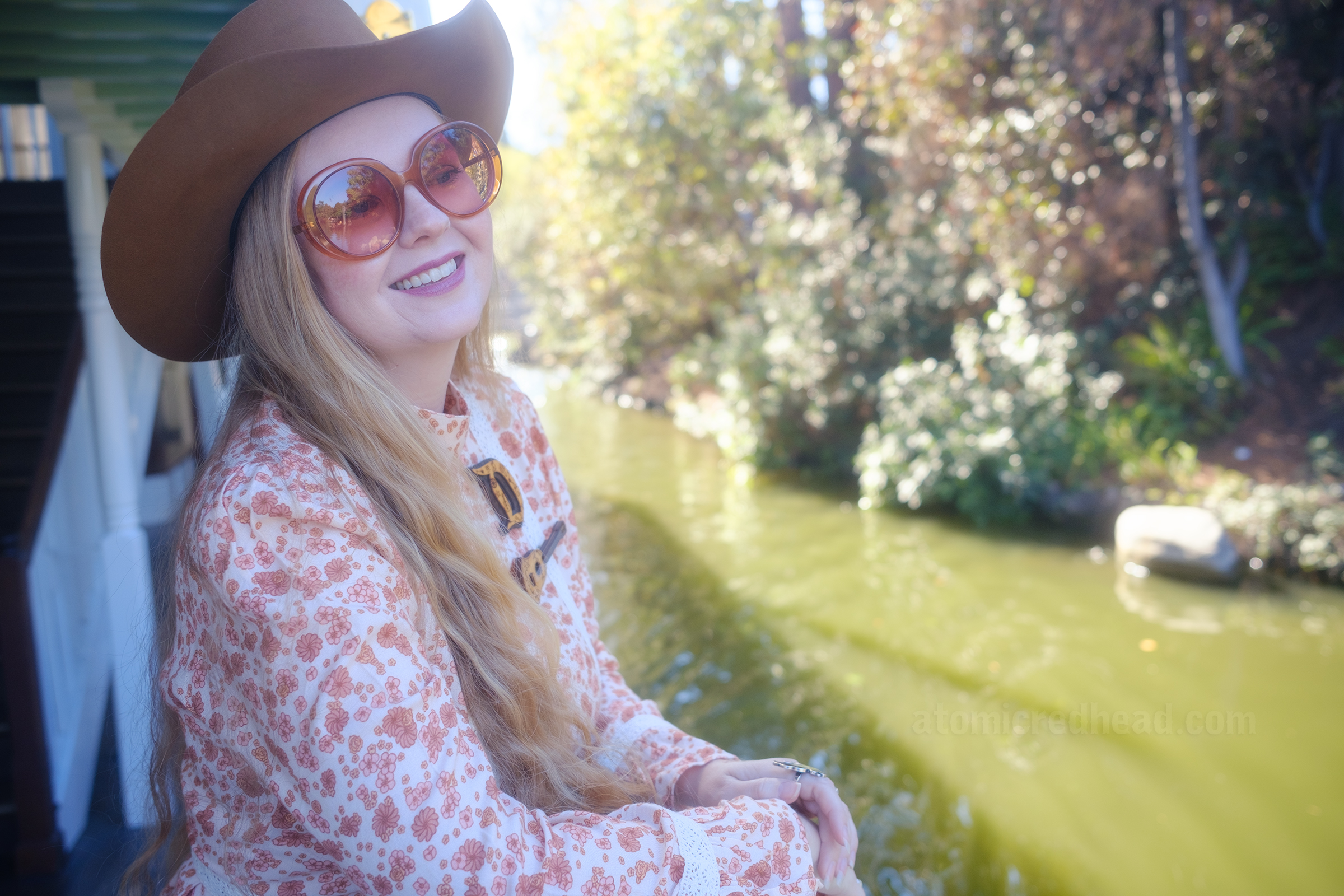 Myself, wearing a light brown cowboy hat, a long sleeve mini dress featuring a floral pattern in shades of orange and brown, with lace detail on the cuffs and waist, and white fringe cowboy boots, leaning against the railing of the Mark Twain Riverboat, looking out at the foliage of the riverbank. 