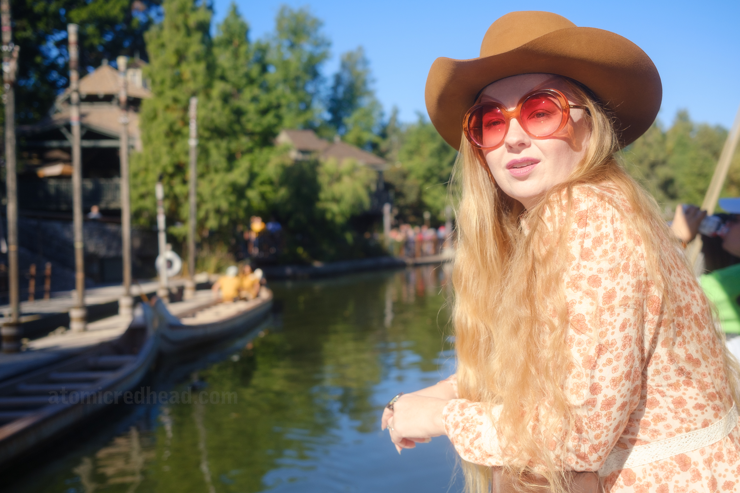 Myself, wearing a light brown cowboy hat, a long sleeve mini dress featuring a floral pattern in shades of orange and brown, with lace detail on the cuffs and waist, and white fringe cowboy boots, leaning against the railing of the Mark Twain Riverboat, looking out at the foliage of the riverbank. 
