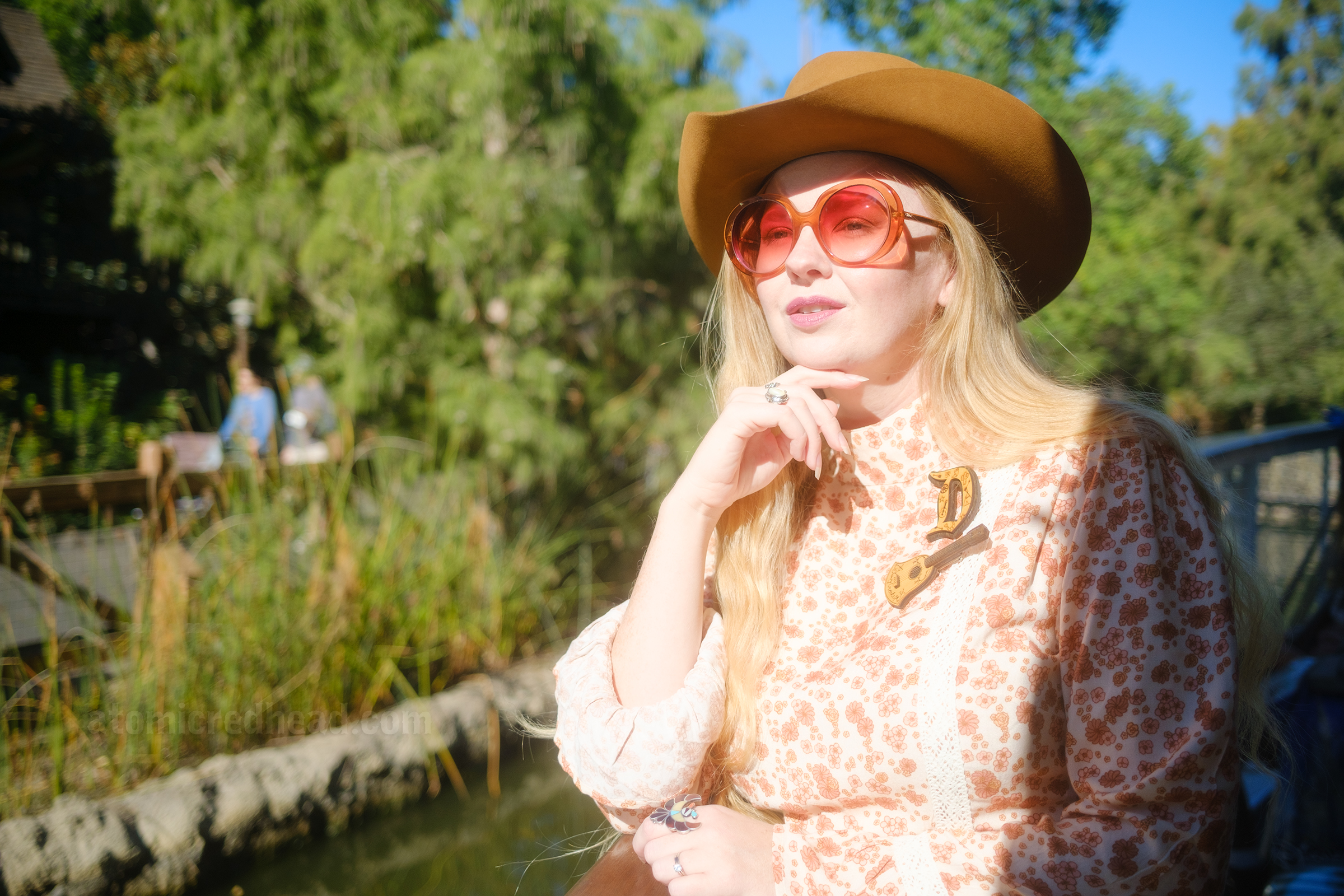 Myself, wearing a light brown cowboy hat, a long sleeve mini dress featuring a floral pattern in shades of orange and brown, with lace detail on the cuffs and waist, and white fringe cowboy boots, leaning against the railing of the Mark Twain Riverboat, looking out at the foliage of the riverbank. 