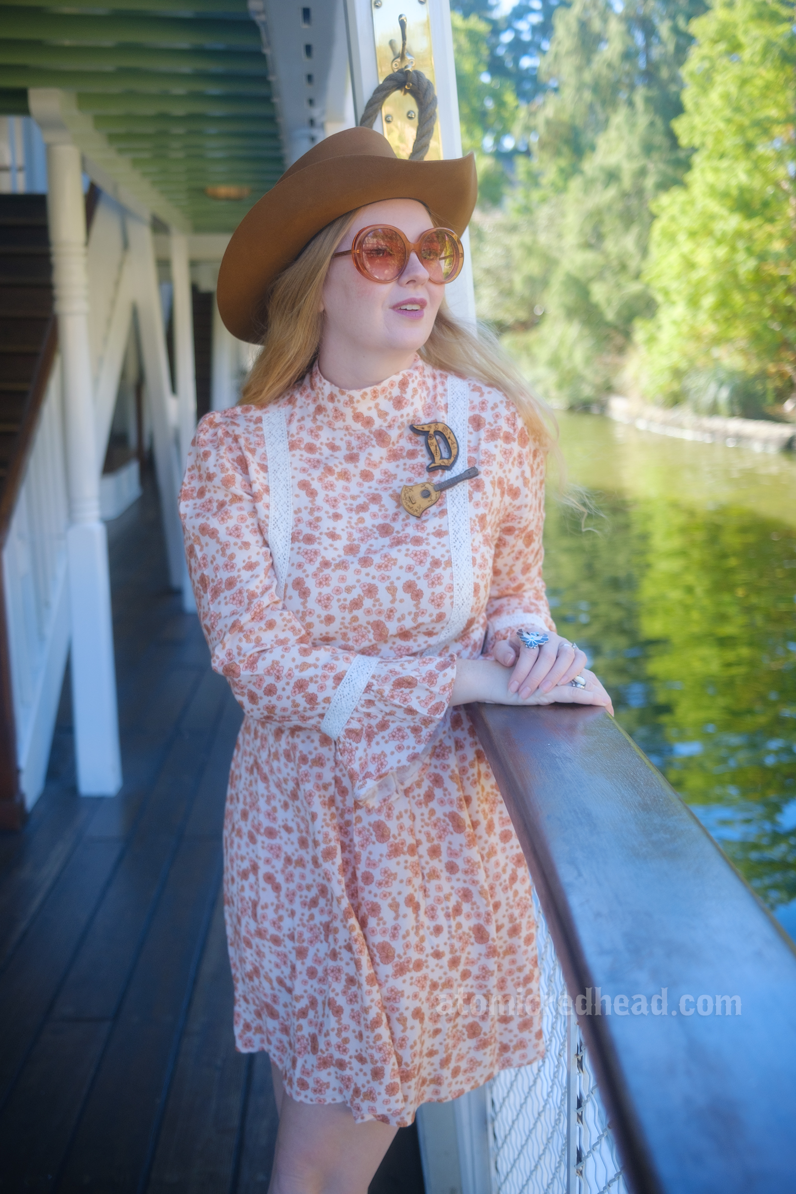 Myself, wearing a light brown cowboy hat, a long sleeve mini dress featuring a floral pattern in shades of orange and brown, with lace detail on the cuffs and waist, and white fringe cowboy boots, leaning against the railing of the Mark Twain Riverboat, looking out at the foliage of the riverbank. 