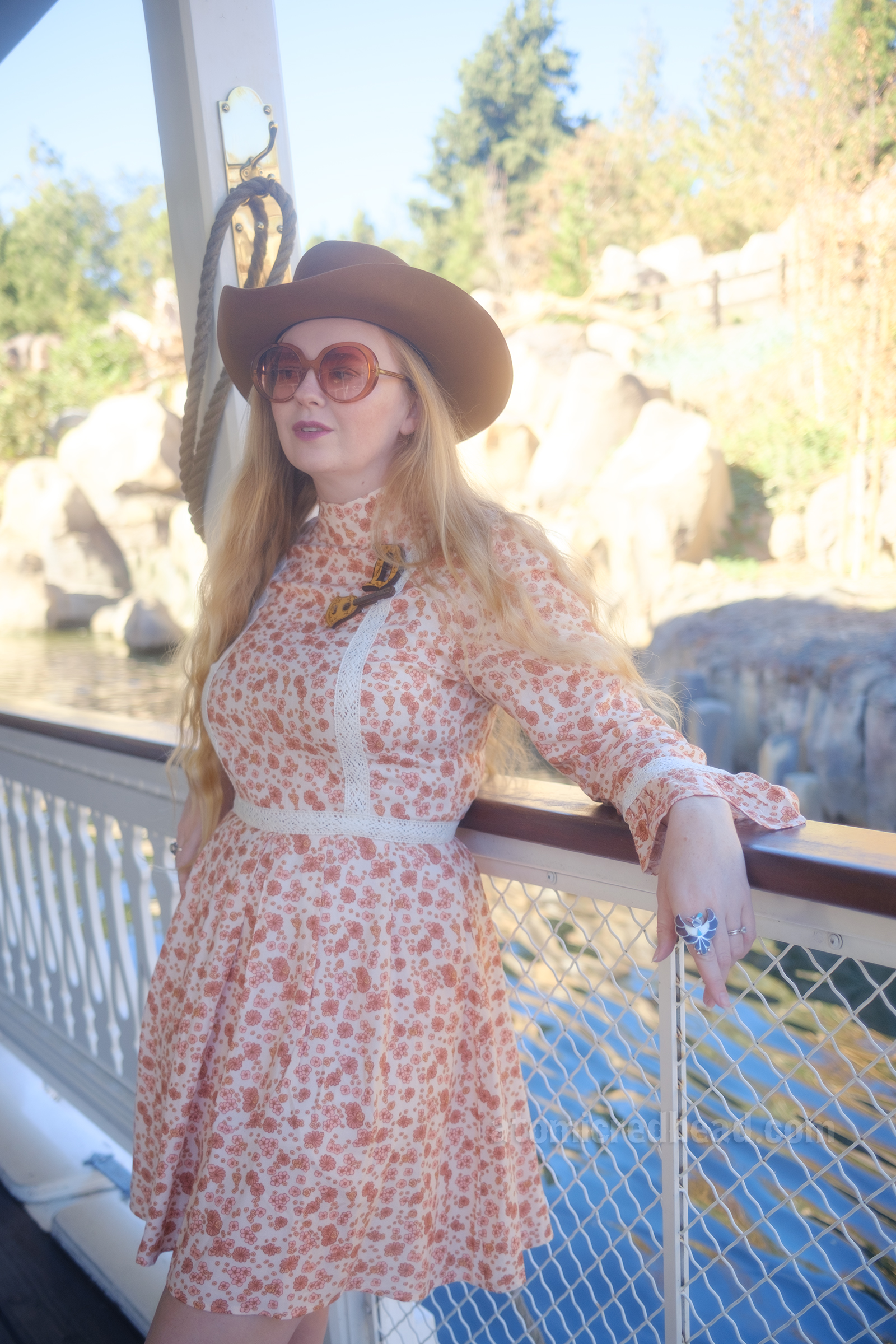 Myself, wearing a light brown cowboy hat, a long sleeve mini dress featuring a floral pattern in shades of orange and brown, with lace detail on the cuffs and waist, and white fringe cowboy boots, leaning against the railing of the Mark Twain Riverboat.
