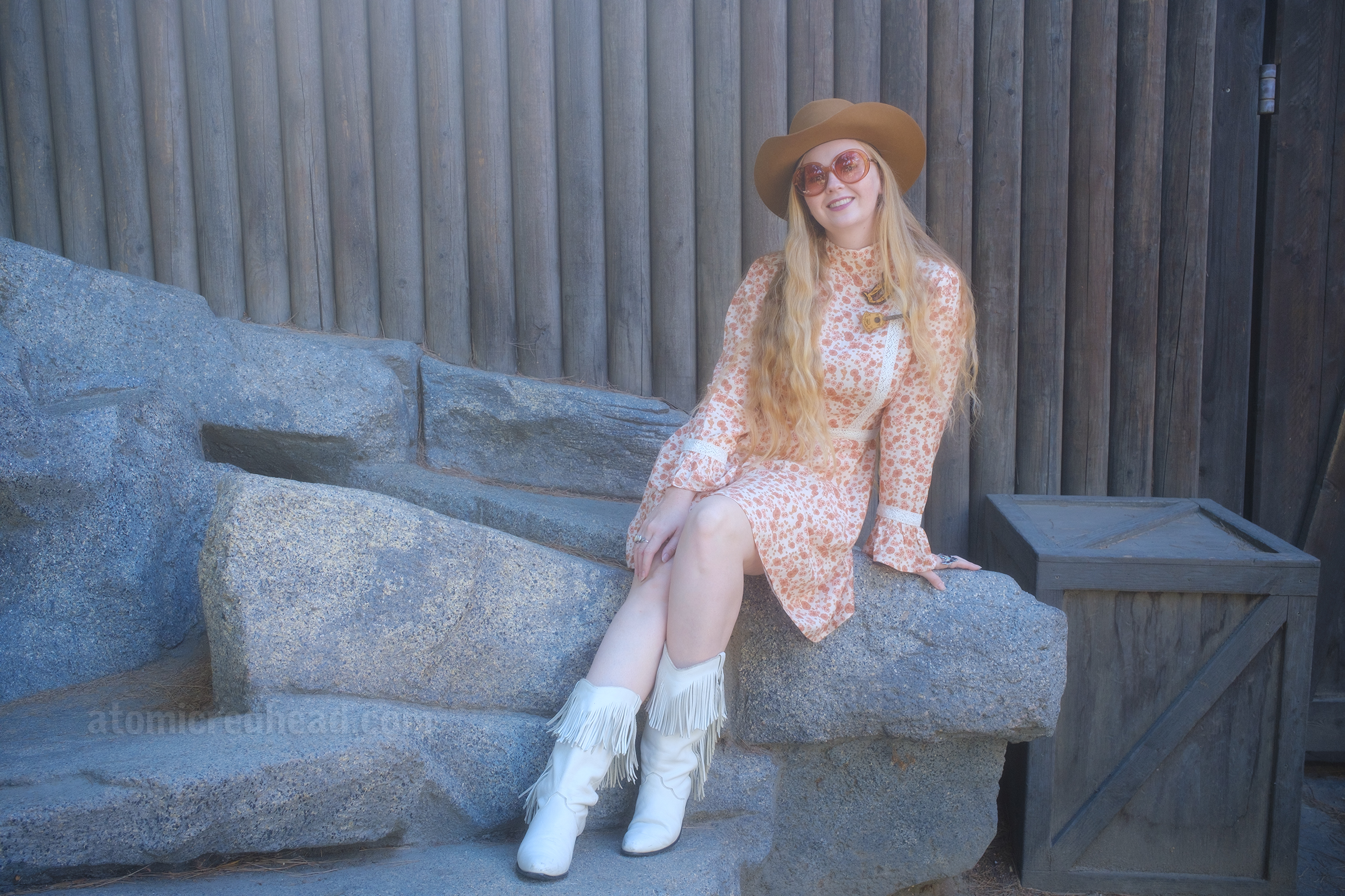 Myself, wearing a light brown cowboy hat, a long sleeve mini dress featuring a floral pattern in shades of orange and brown, with lace detail on the cuffs and waist, and white fringe cowboy boots, sitting on a rock that is up against the wooden Fort Wilderness on Tom Sawyer's Island.