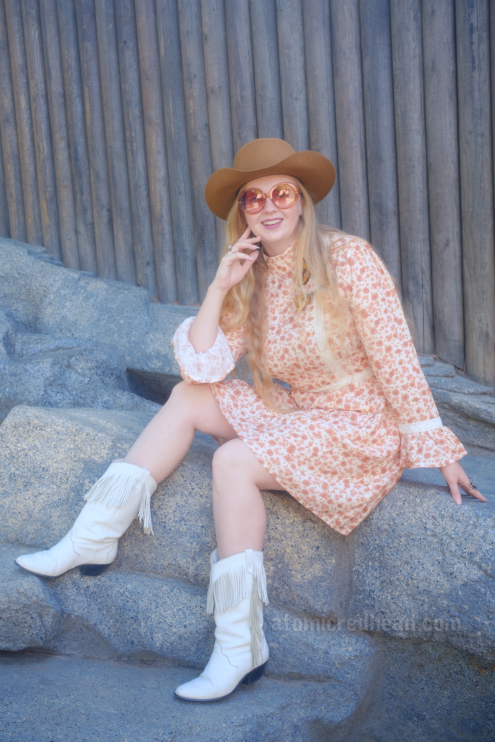 Myself, wearing a light brown cowboy hat, a long sleeve mini dress featuring a floral pattern in shades of orange and brown, with lace detail on the cuffs and waist, and white fringe cowboy boots, sitting on a rock that is up against the wooden Fort Wilderness on Tom Sawyer's Island.