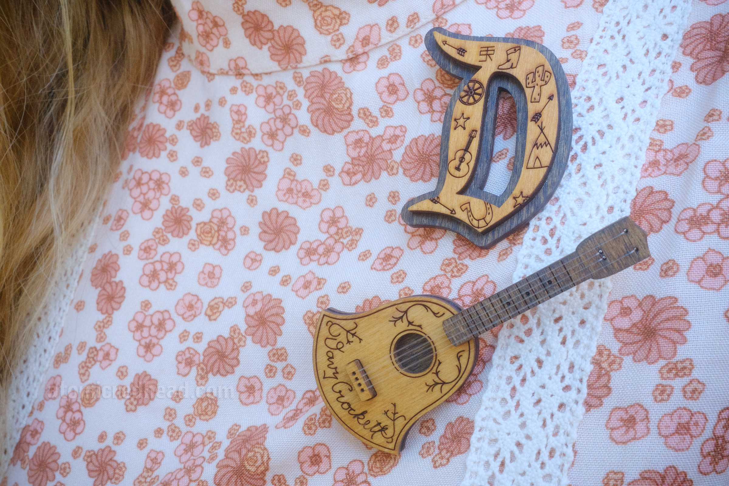 Close-up of my brooches, one is a D (in the style of the iconic, gothic Disneyland D) filled with icons of Frontierland, such as a cactus, star, arrow, and boot, the other is a miniature guitar that reads "Davy Crockett" across the bottom of it.