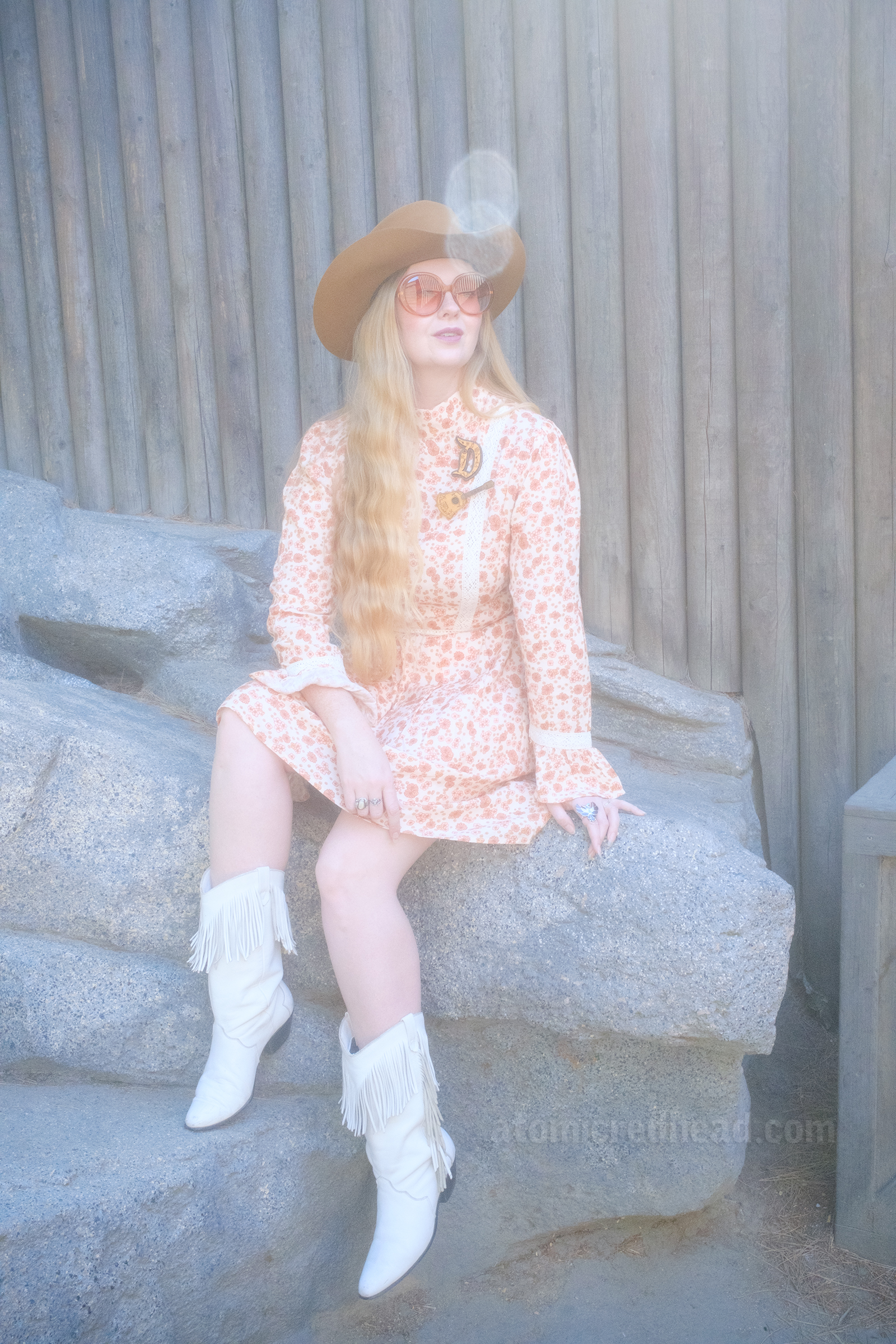 Myself, wearing a light brown cowboy hat, a long sleeve mini dress featuring a floral pattern in shades of orange and brown, with lace detail on the cuffs and waist, and white fringe cowboy boots, sitting on a rock that is up against the wooden Fort Wilderness on Tom Sawyer's Island.