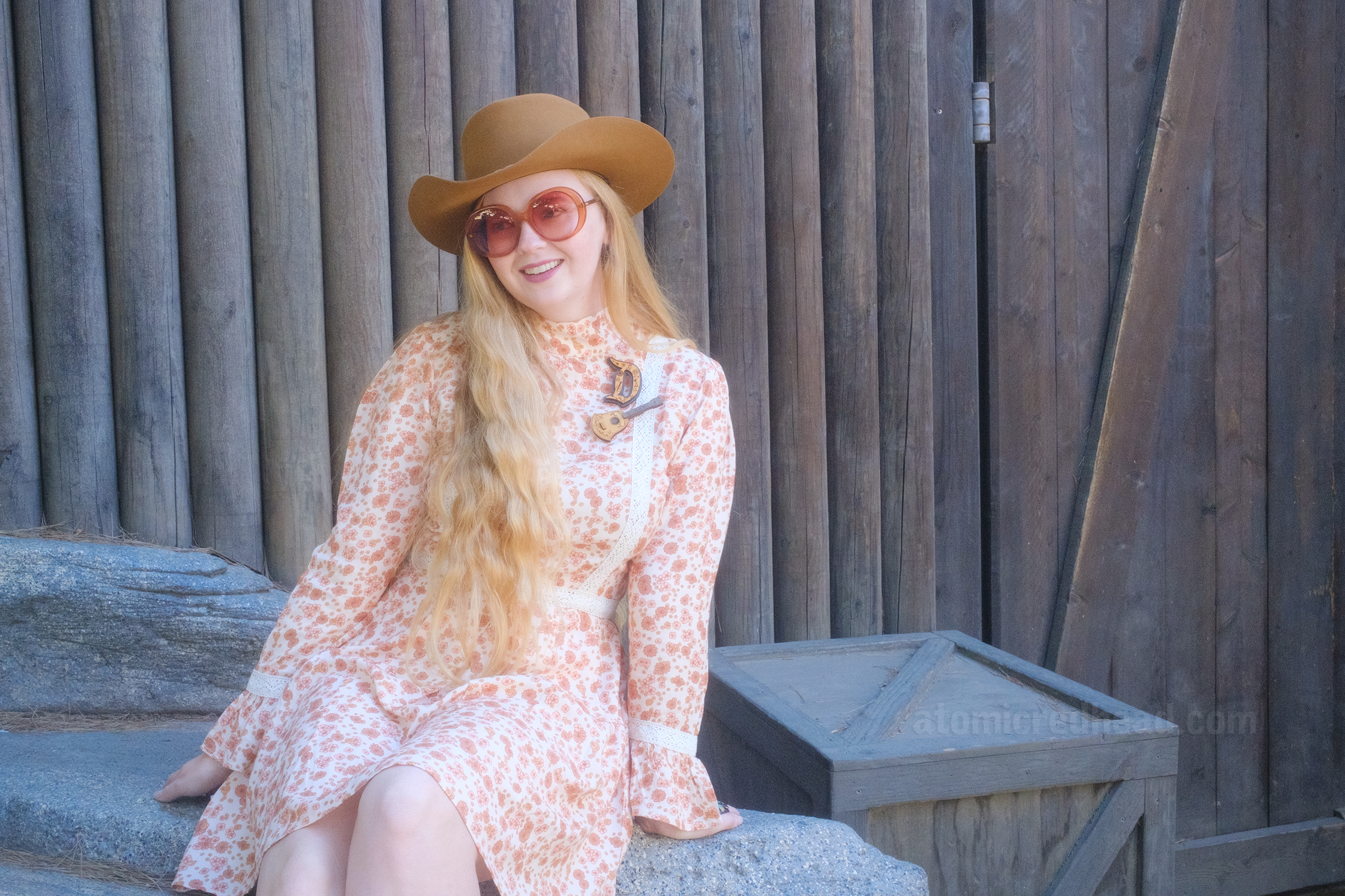 Myself, wearing a light brown cowboy hat, a long sleeve mini dress featuring a floral pattern in shades of orange and brown, with lace detail on the cuffs and waist, and white fringe cowboy boots, sitting on a rock that is up against the wooden Fort Wilderness on Tom Sawyer's Island.