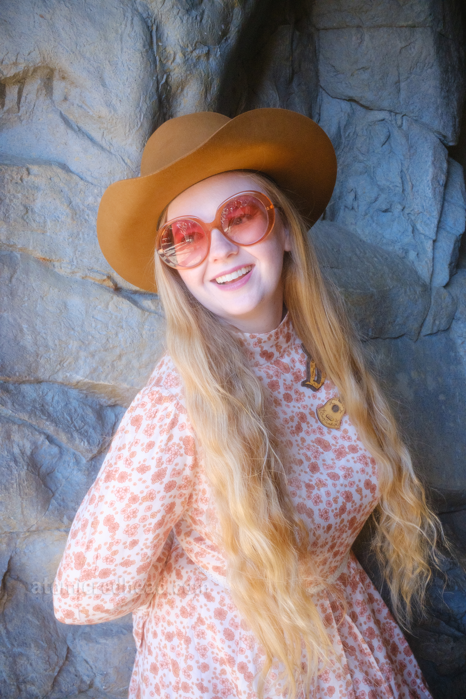 Myself, wearing a light brown cowboy hat, a long sleeve mini dress featuring a floral pattern in shades of orange and brown, with lace detail on the cuffs and waist, and white fringe cowboy boots, leaning against a rock wall.