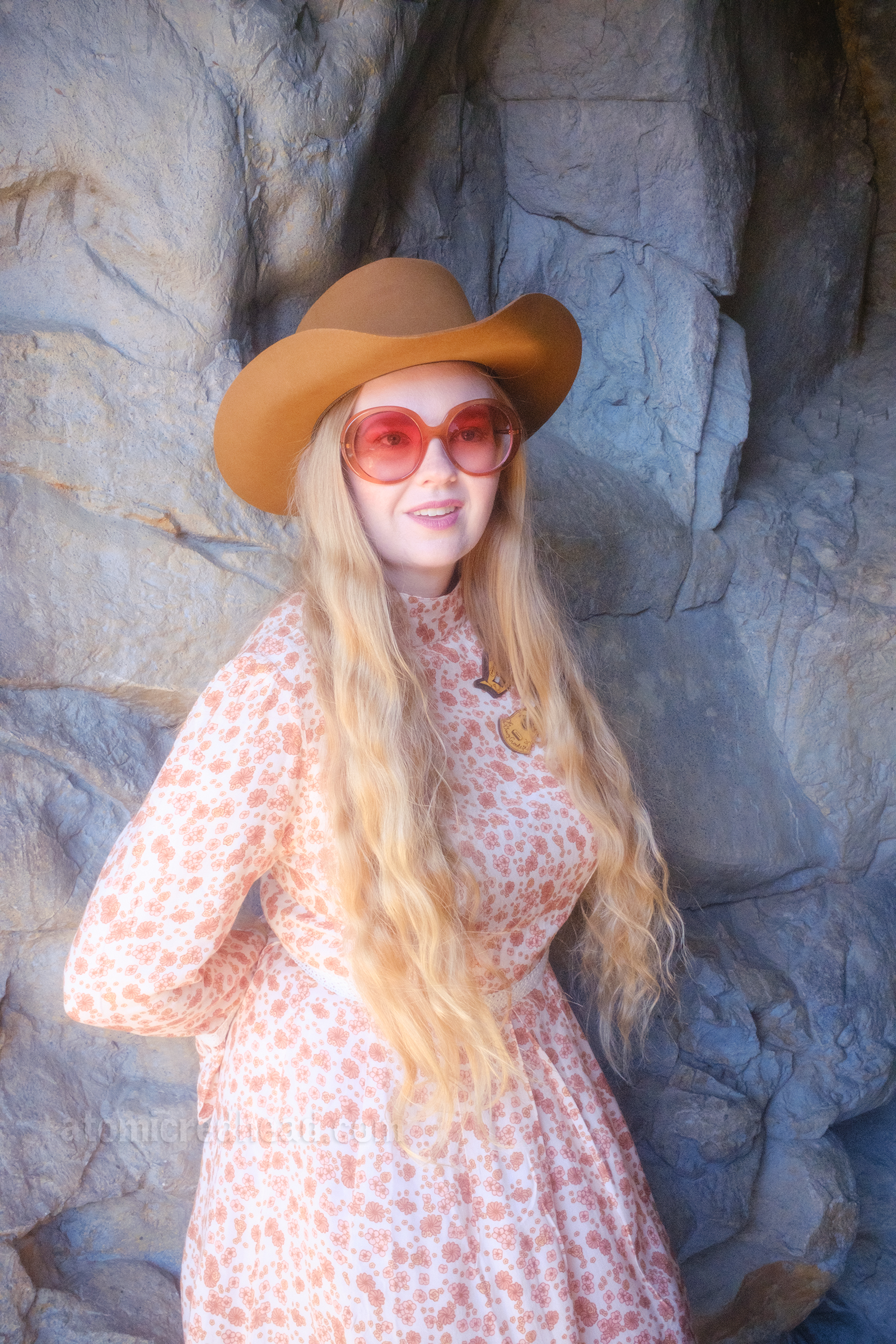 Myself, wearing a light brown cowboy hat, a long sleeve mini dress featuring a floral pattern in shades of orange and brown, with lace detail on the cuffs and waist, and white fringe cowboy boots, leaning against a rock wall.