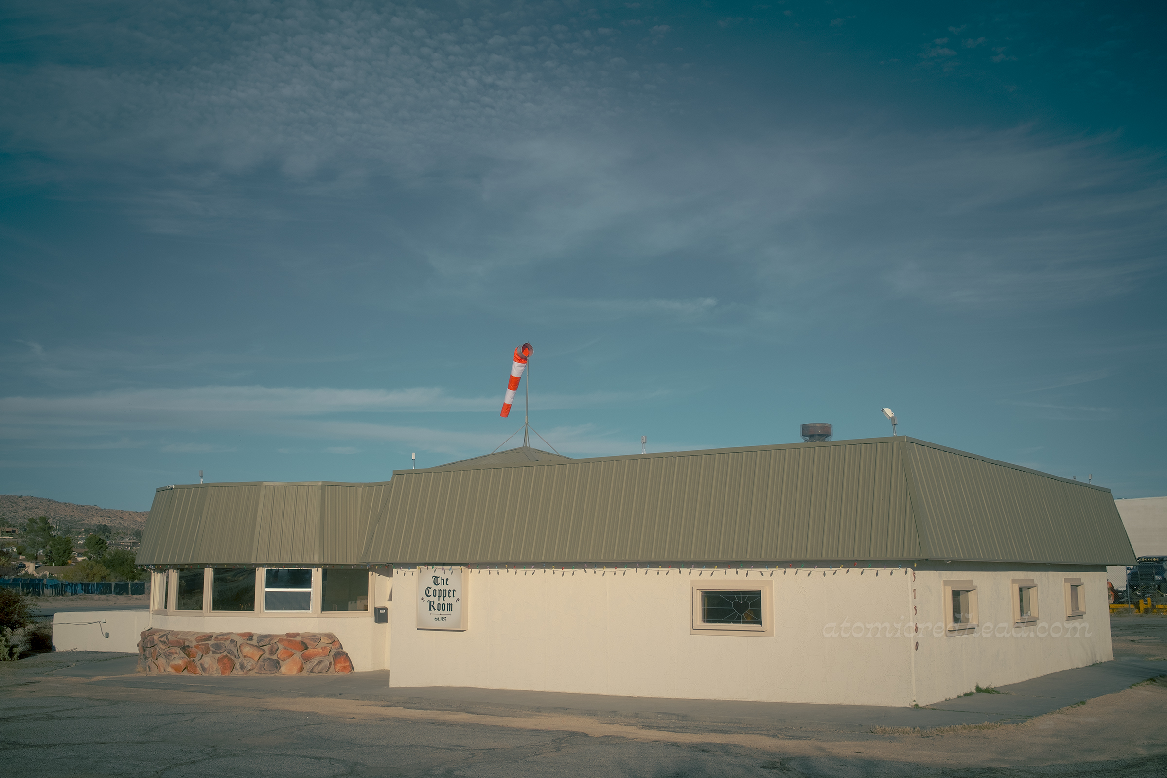 A small white stucco building with a green roof, a curved portion features warm rocks along the wall, a small sign reads "The Copper Room est. 1957." Atop the roof is a small orange wind sock.