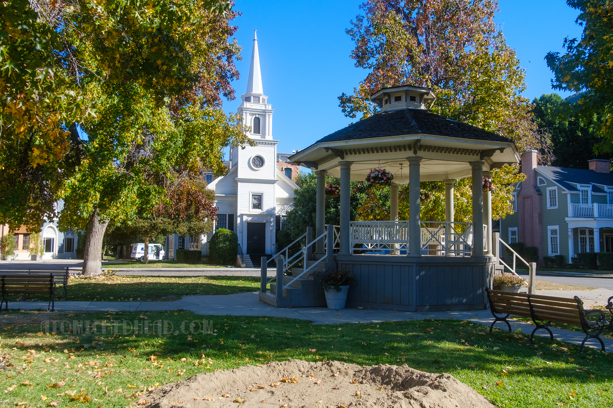 A small gazebo rests in a grassy square as part of Midwest Street, in the background a white steeple church is visible.