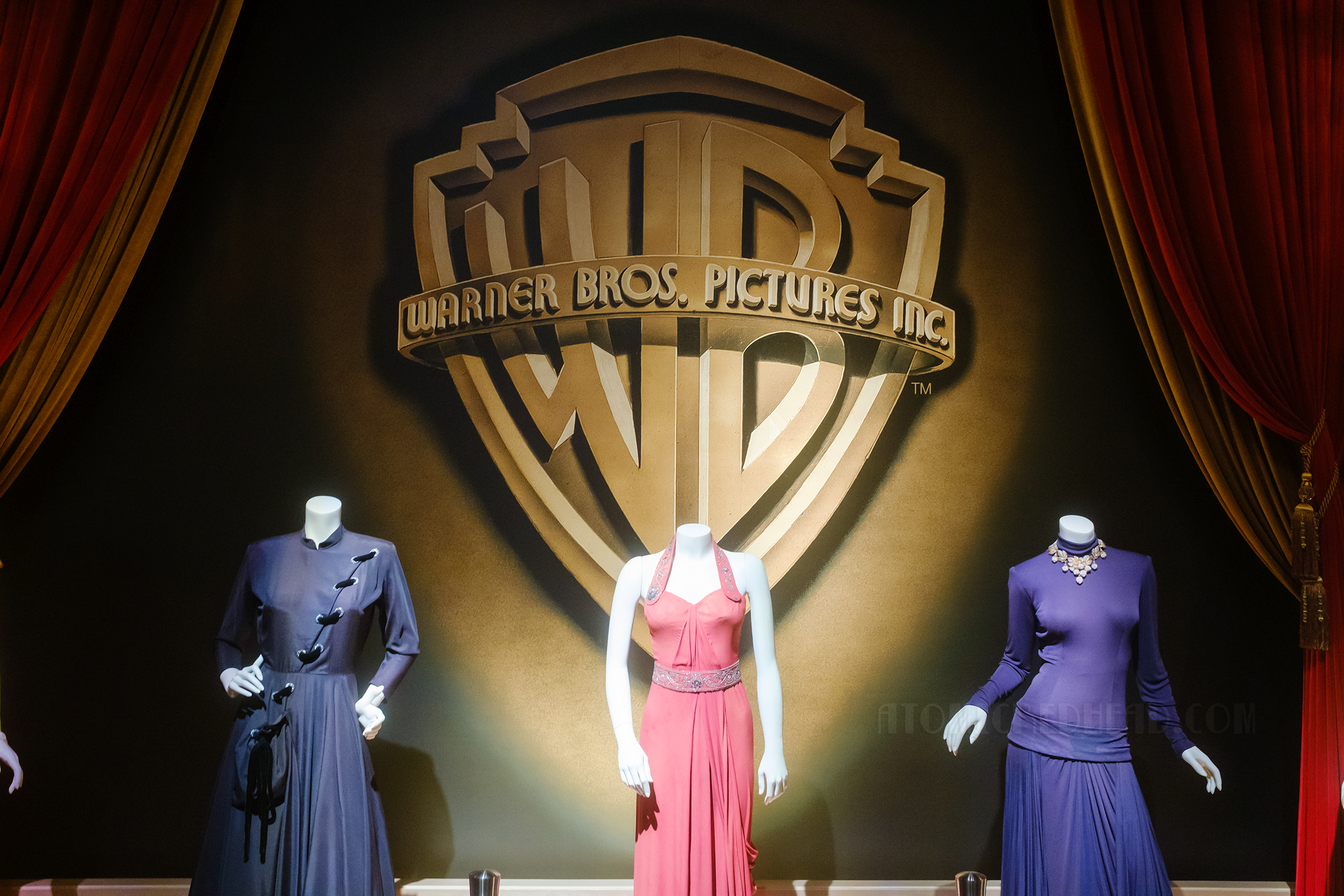 The large golden Warner Bros. logo rises above three mannequins which wear dresses, the left is a blue dress with velvet detail, the middle a pink halter dress with beading at the waist, and a drop waist long sleeve navy dress on the right.