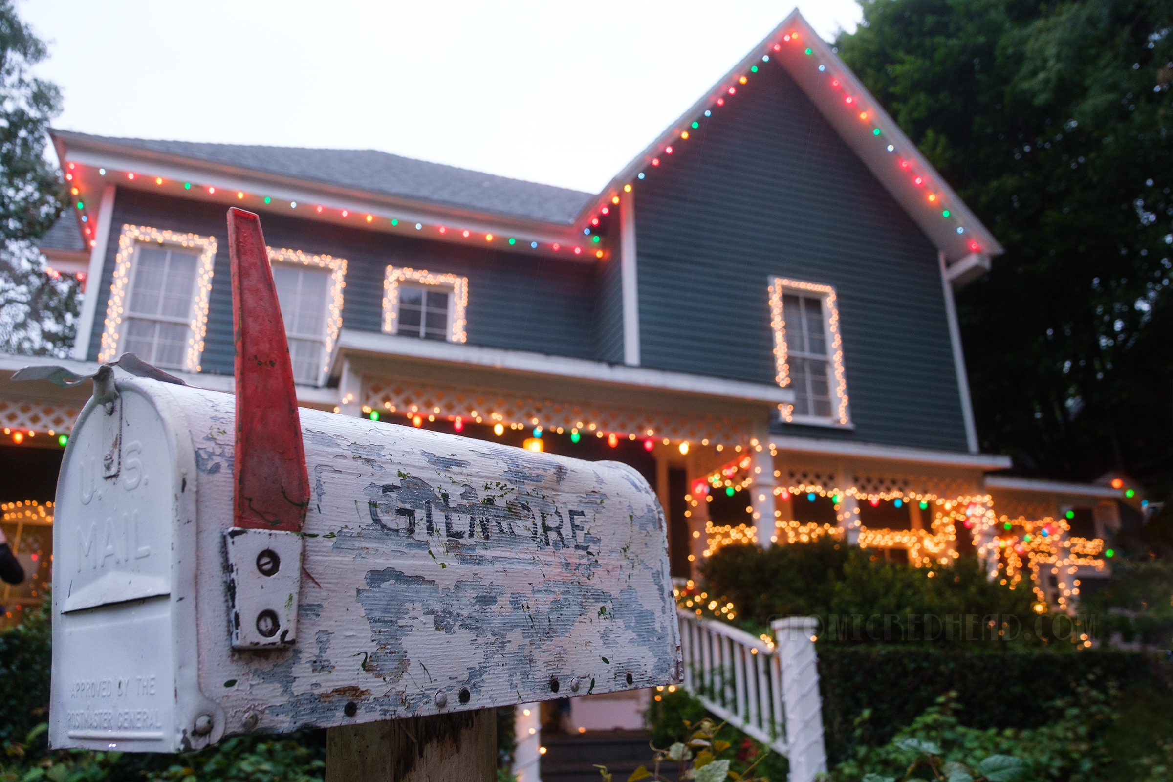 The Gilmore Girls home, a green and white farmhouse style home with Christmas lights, and a white mail box outside with "Gilmore" painted on the side.