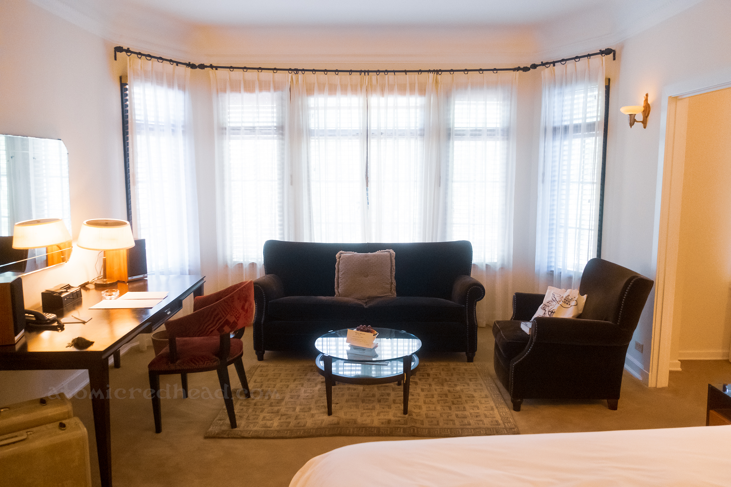 Overall view of the living space of our room, which features a dark wood desk on the left with a chair, a dark upholstered couch, and matching chair, with a circular table in the middle.