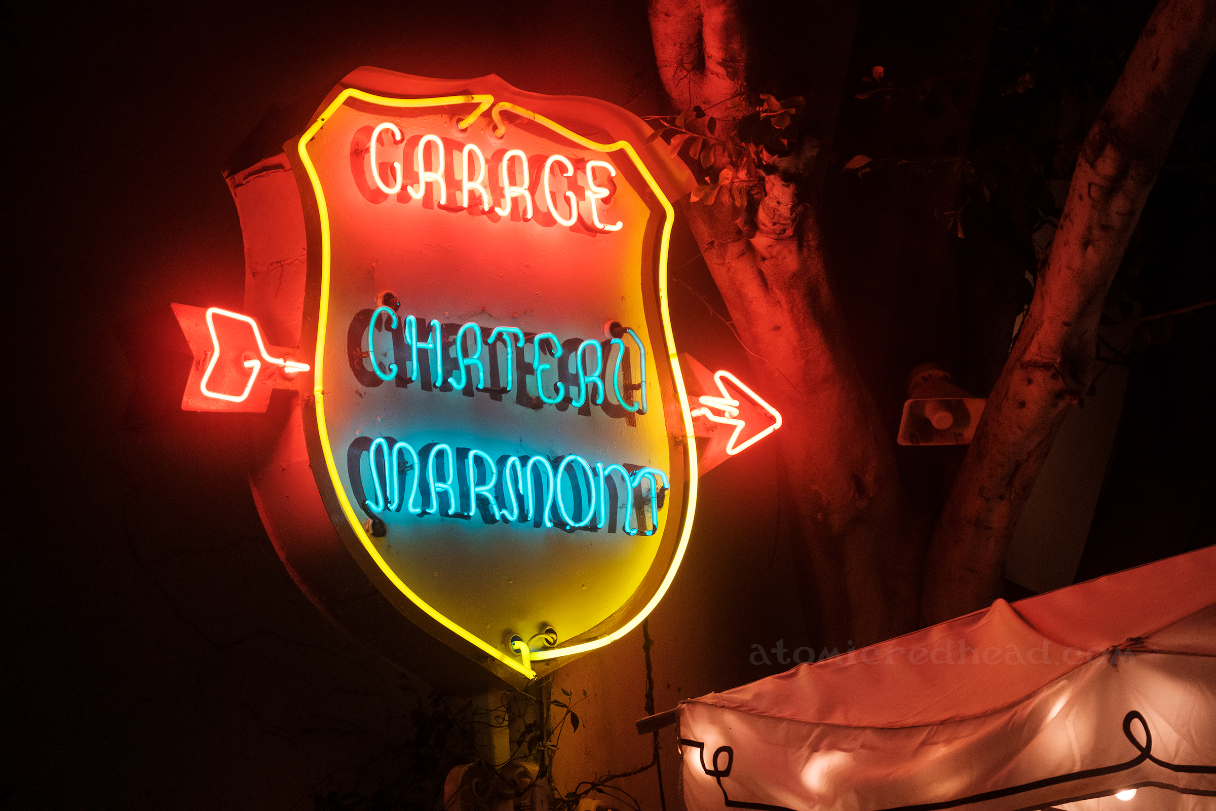 A shield shaped neon sign has a red neon arrow passing through it. Red neon letters read "Garage" and below in blue neon letters reads "Chateau Marmont"