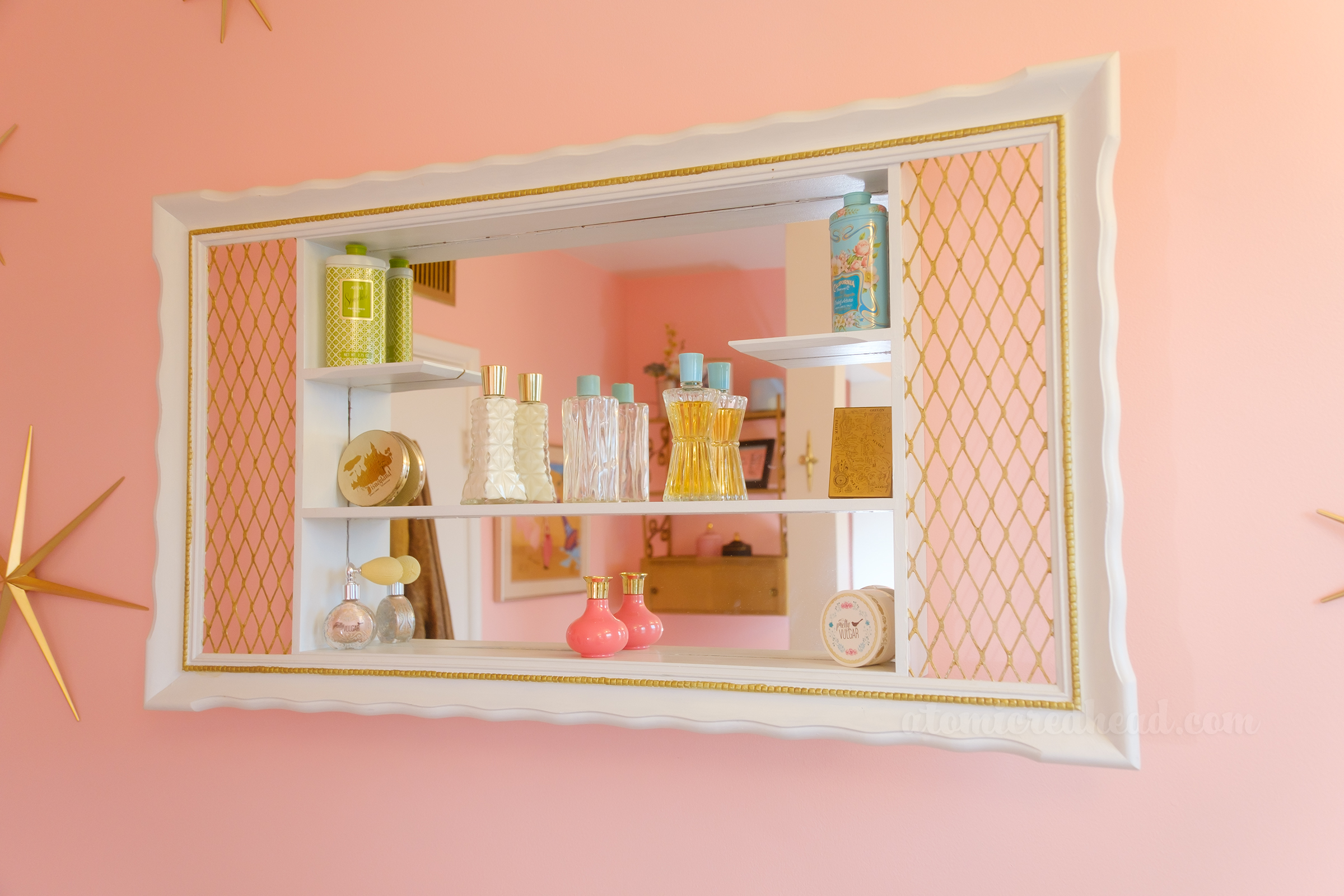 A white and gold shadow box hangs on a pink wall. Resting within the shadow box are a variety of vintage bottles and compacts.