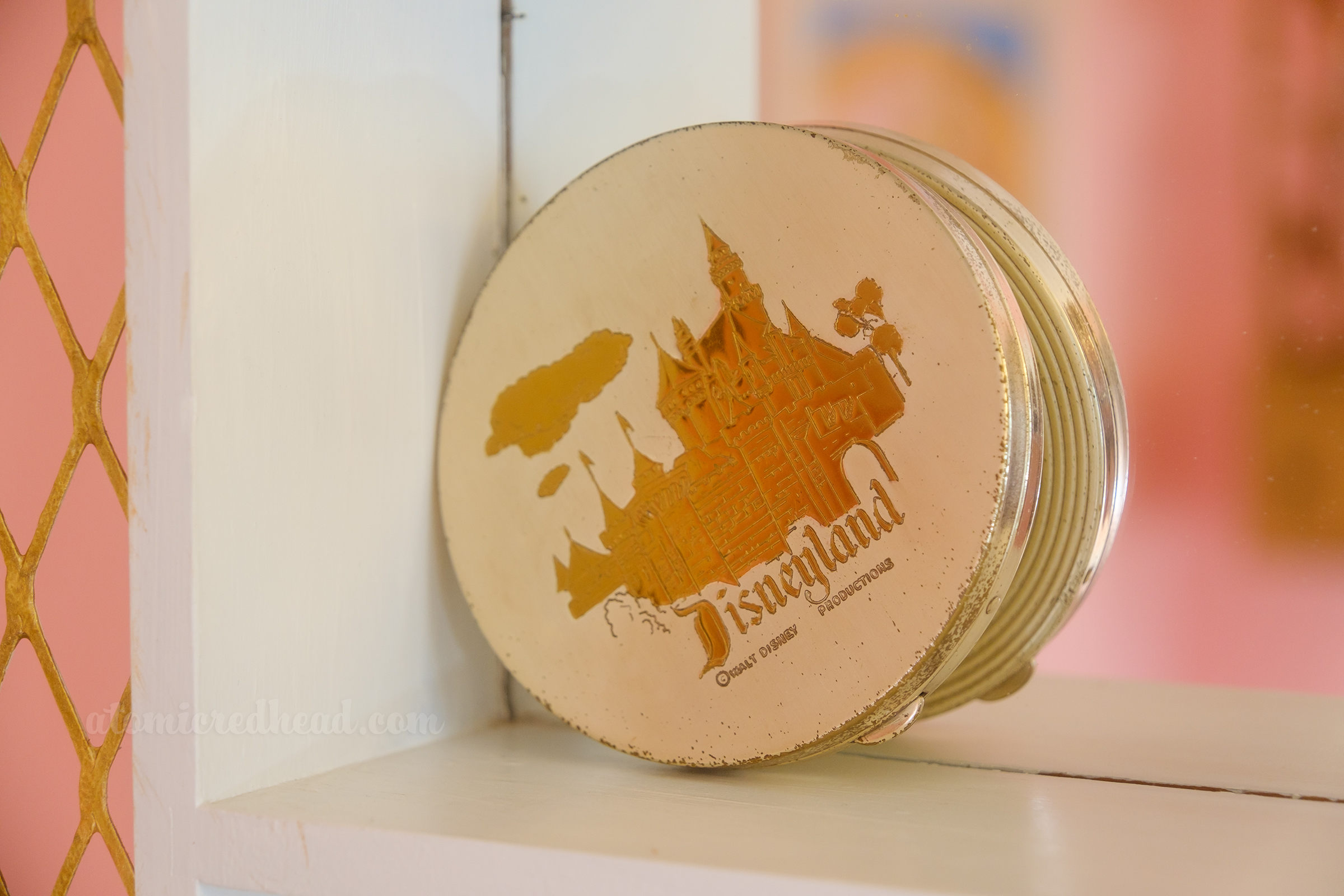 A round gold tone compact featuring Sleeping Beauty Castle and text reading "Disneyland" rests on a shelf inside the shadowbox.