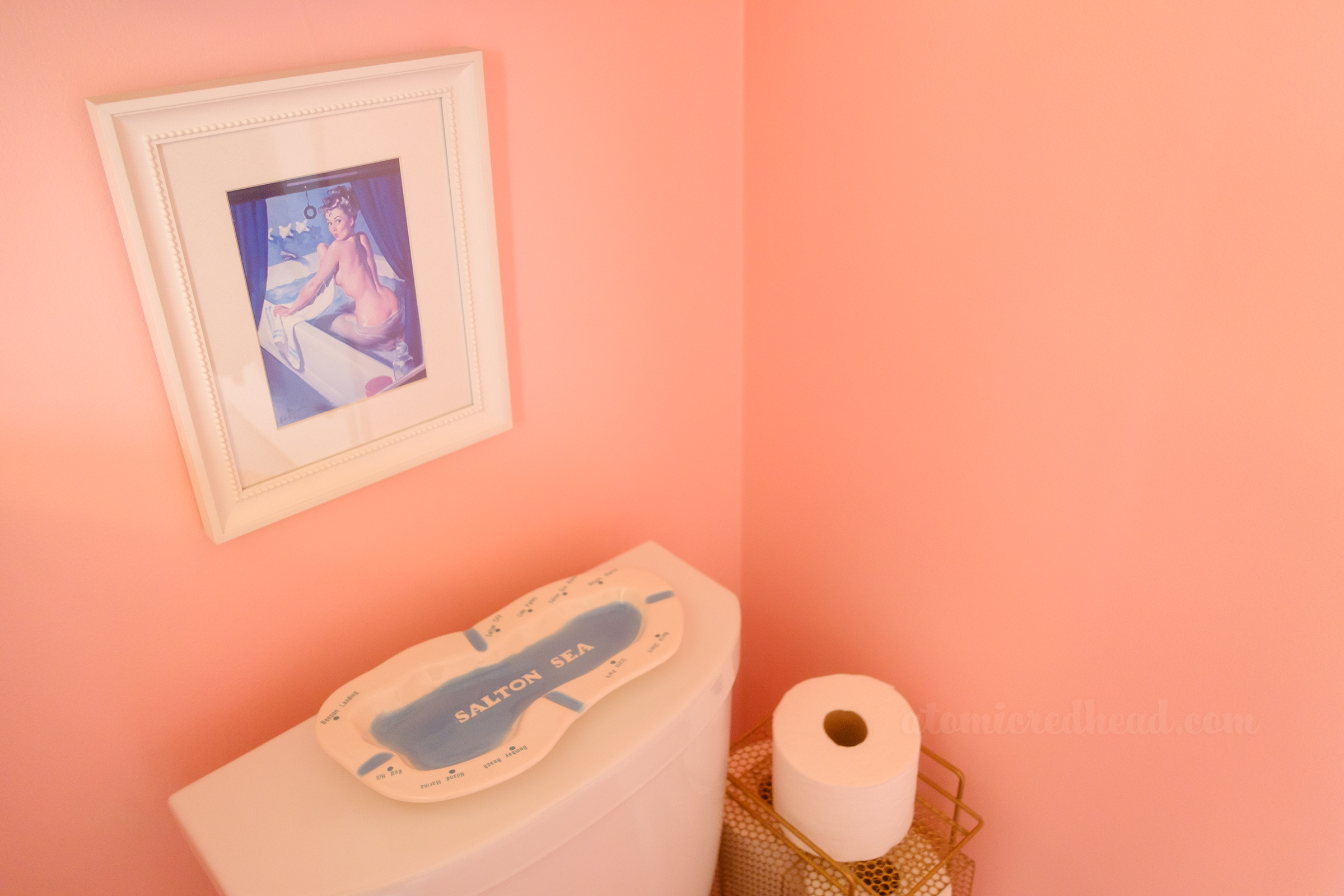 A framed image of a pin-up rising from her bathtub hangs above the toilet. Resting on top of the toilet tank is an ashtray in the shape of and reading "Salton Sea" with dots noting the various towns around the Salton Sea.