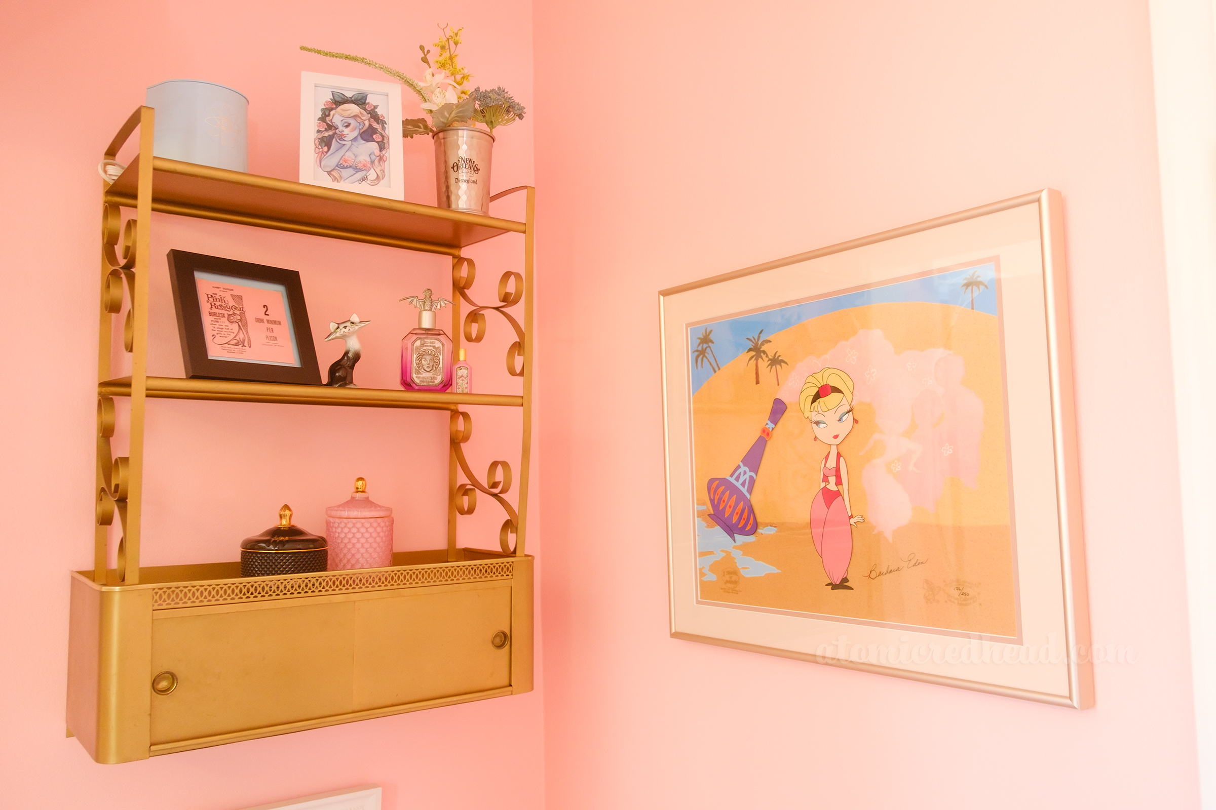 The area above the toilet, on the left is a gold shelf with various bottles and framed works of art. To the right is a large framed cel of the animation sequence of the opening of I Dream of Jeannie, featuring Jeannie, in her pink harem girl costume, and her purple bottle.
