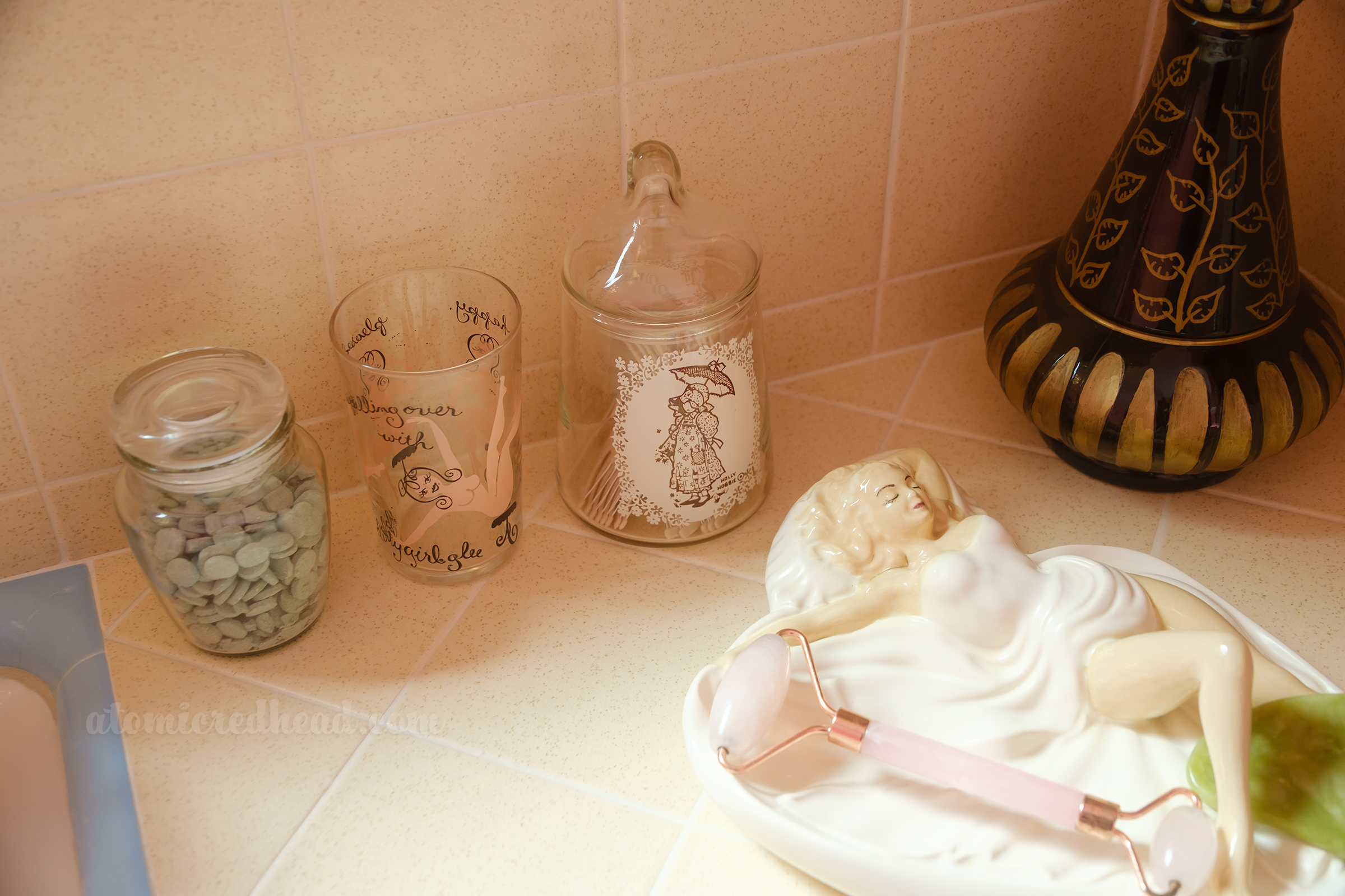 A small glass jar holds green mouthwash tablets, a glass rests next to that featuring a naked cartoon woman, and next to that a small glass jar featuring Holly Hobby on it holds floss picks. To the right is a dish of a woman laying down, which holds a rose quartz roller.