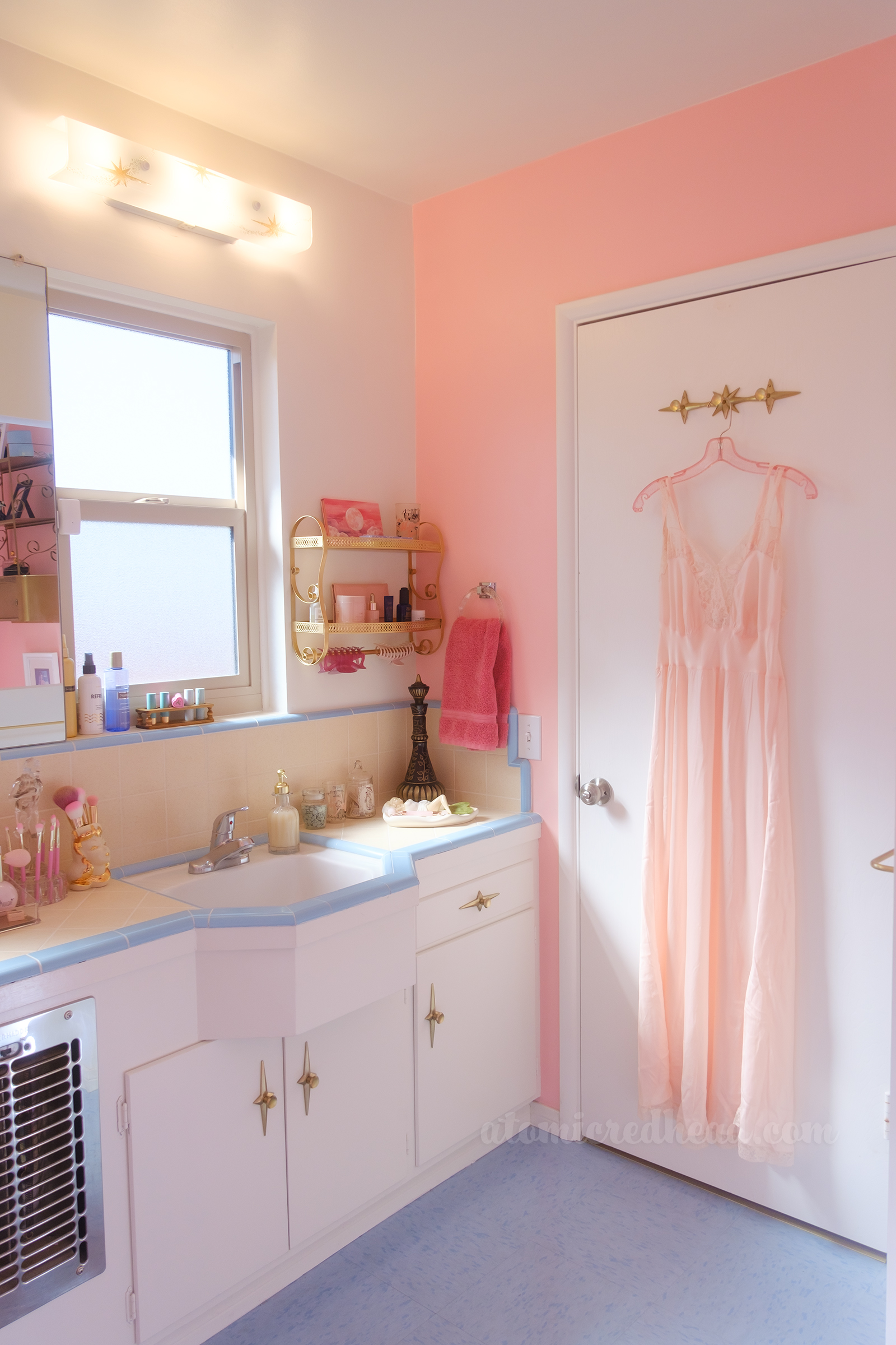 Pink walls meet a yellow and blue tile countertop that has several bits and bobbles on it. On the back of the door is a trio of gold starburst hooks, on one hangs a pink glitter hanger holding a pink slip.