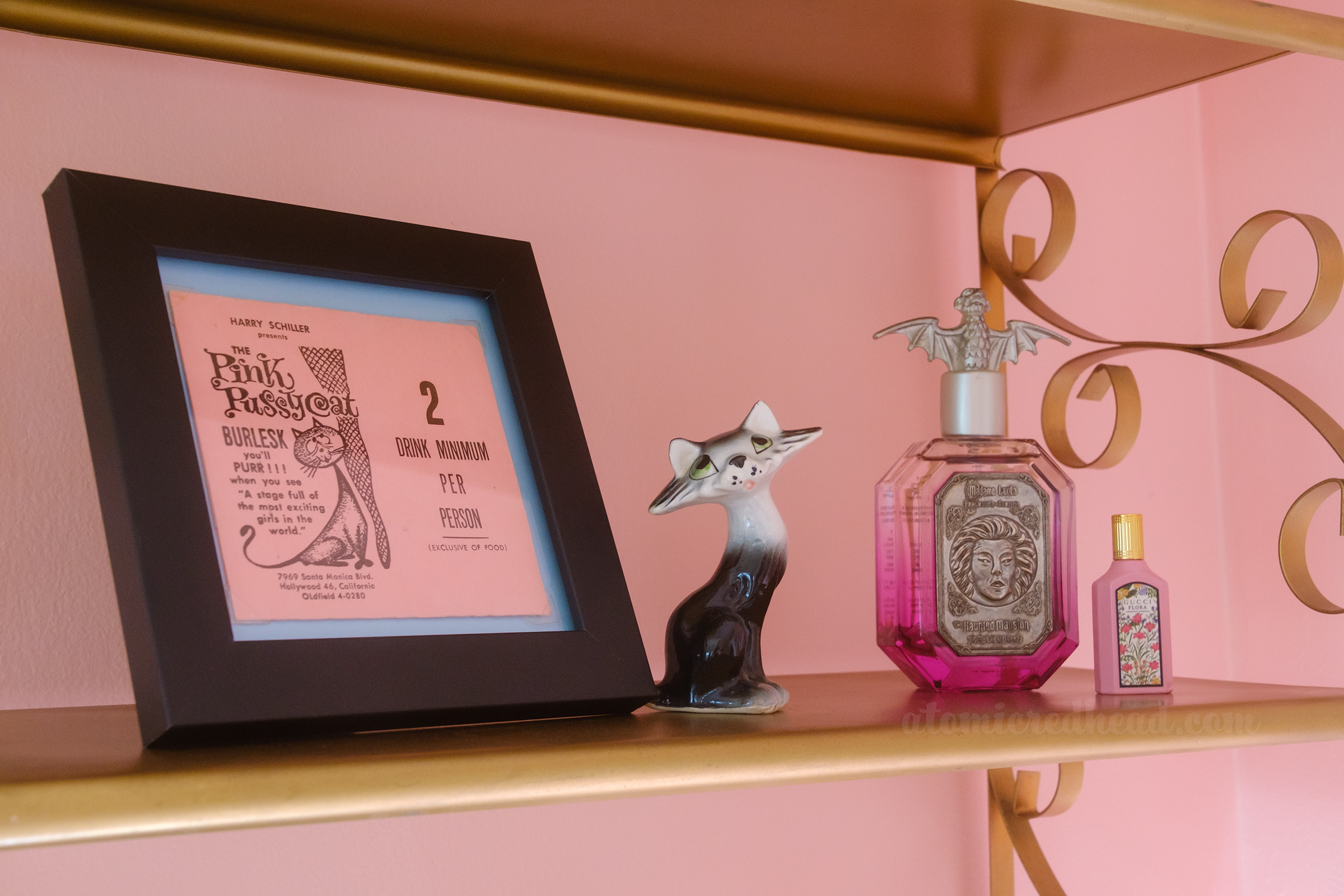 Close-up of a framed table placard from the Pink Pussycat, featuring the image of a cat rubbing up against a leg with fishnet stockings on it, text reads "Harry Shiller presents the Pink Pussycat Burlesk you'll purr!! when you see 'a stagefull of the most exciting girls in the world' 2 Drink Minimum" To the right is a small ceramic figure of a grey cat, and next to it is a perfume bottle.