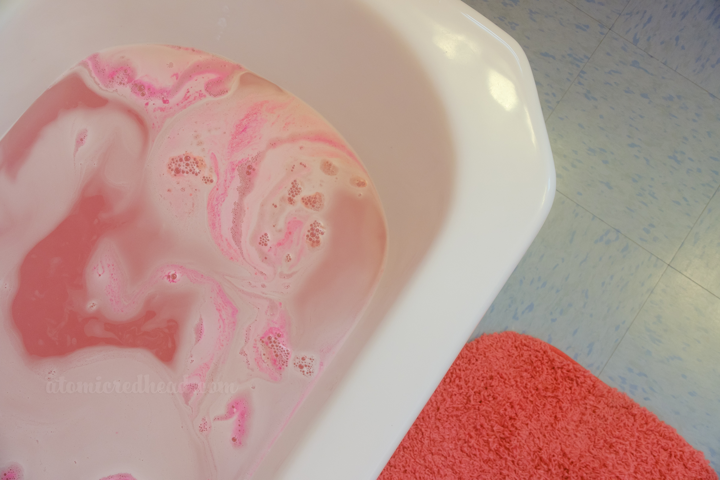 A tub fills with water, a pink bath bomb swirls inside. Outside of the tub, a pink rug resting on blue flooring.