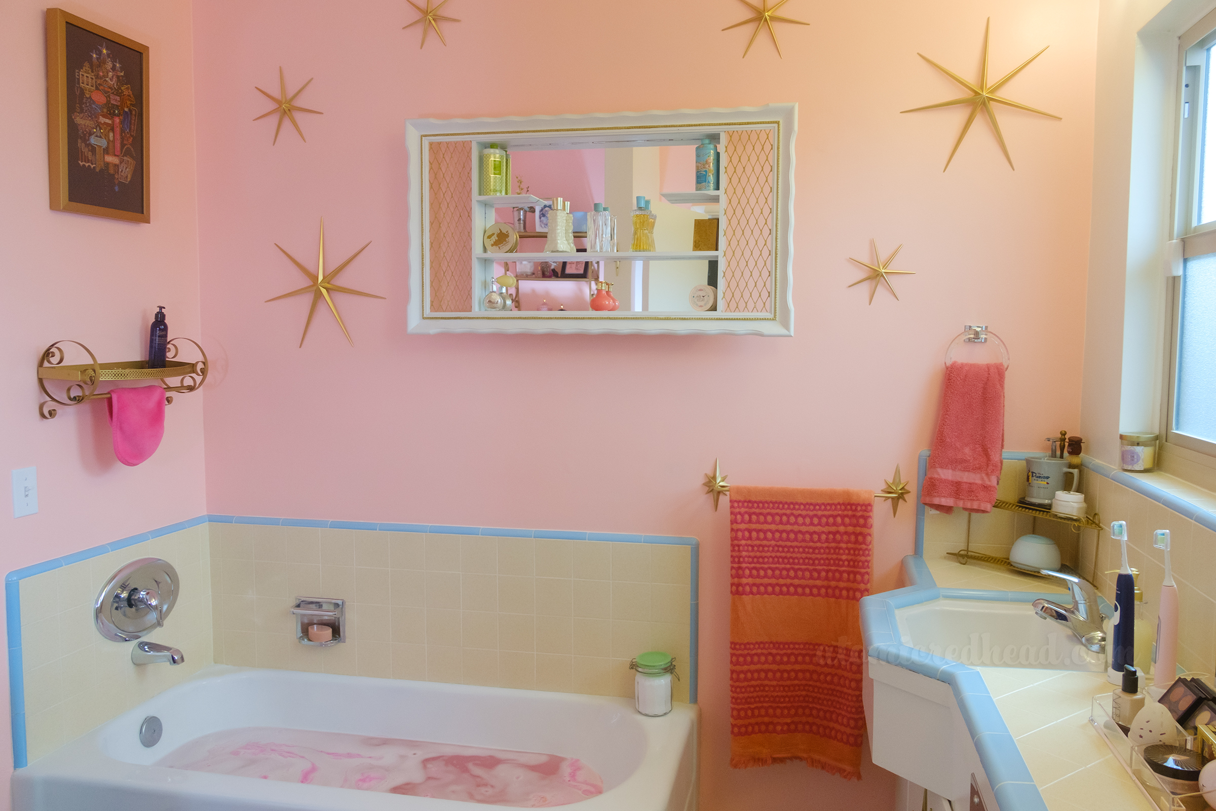 View of the bathtub, which sits in the corner. Hanging above one part is a print featuring vintage Las Vegas signs, above another portion is a large white and gold mirrored shadowbox with various vintage bottles and compacts on it, surrounding the shadow box are a variety of gold starbursts made of metal. To the right of the tub is a gold towel bar with an orange and pink towel hanging on it.