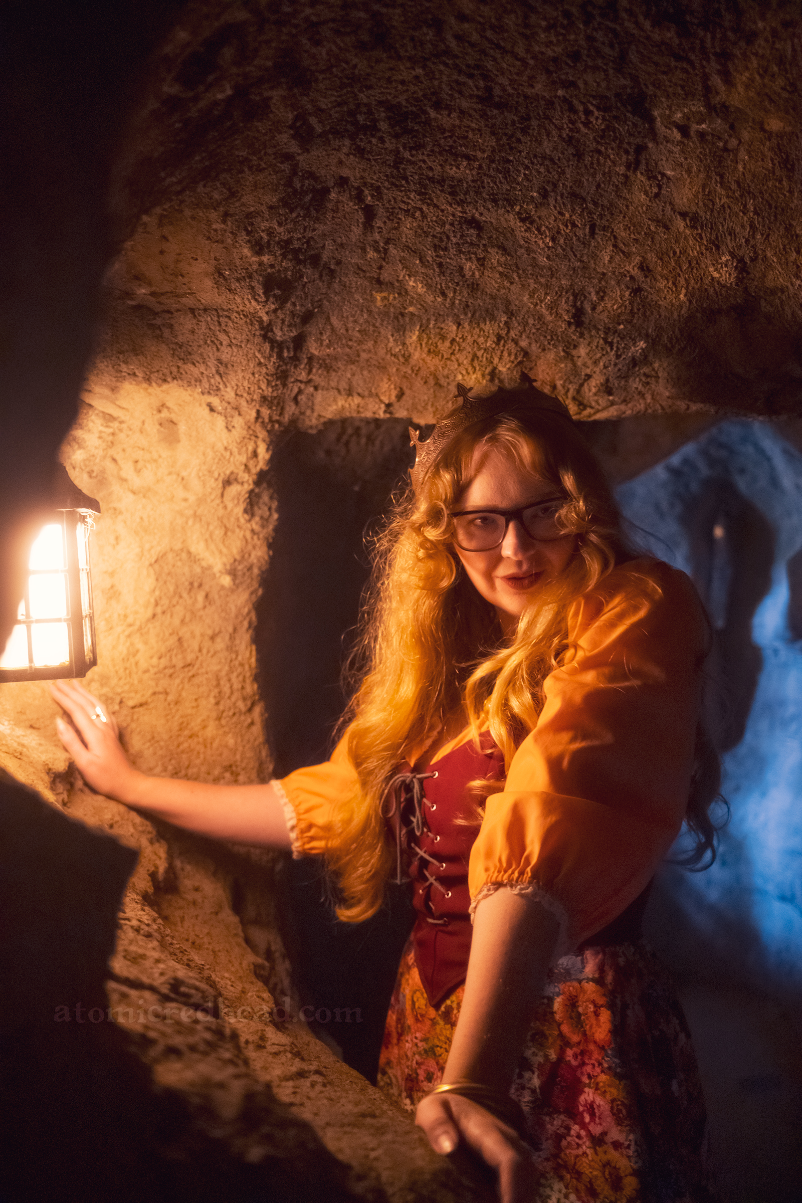 Myself standing in a cave with a light on the left, wearing a copper crown, dress with coral sleeves, maroon lace up bodice, and short floral skirt.