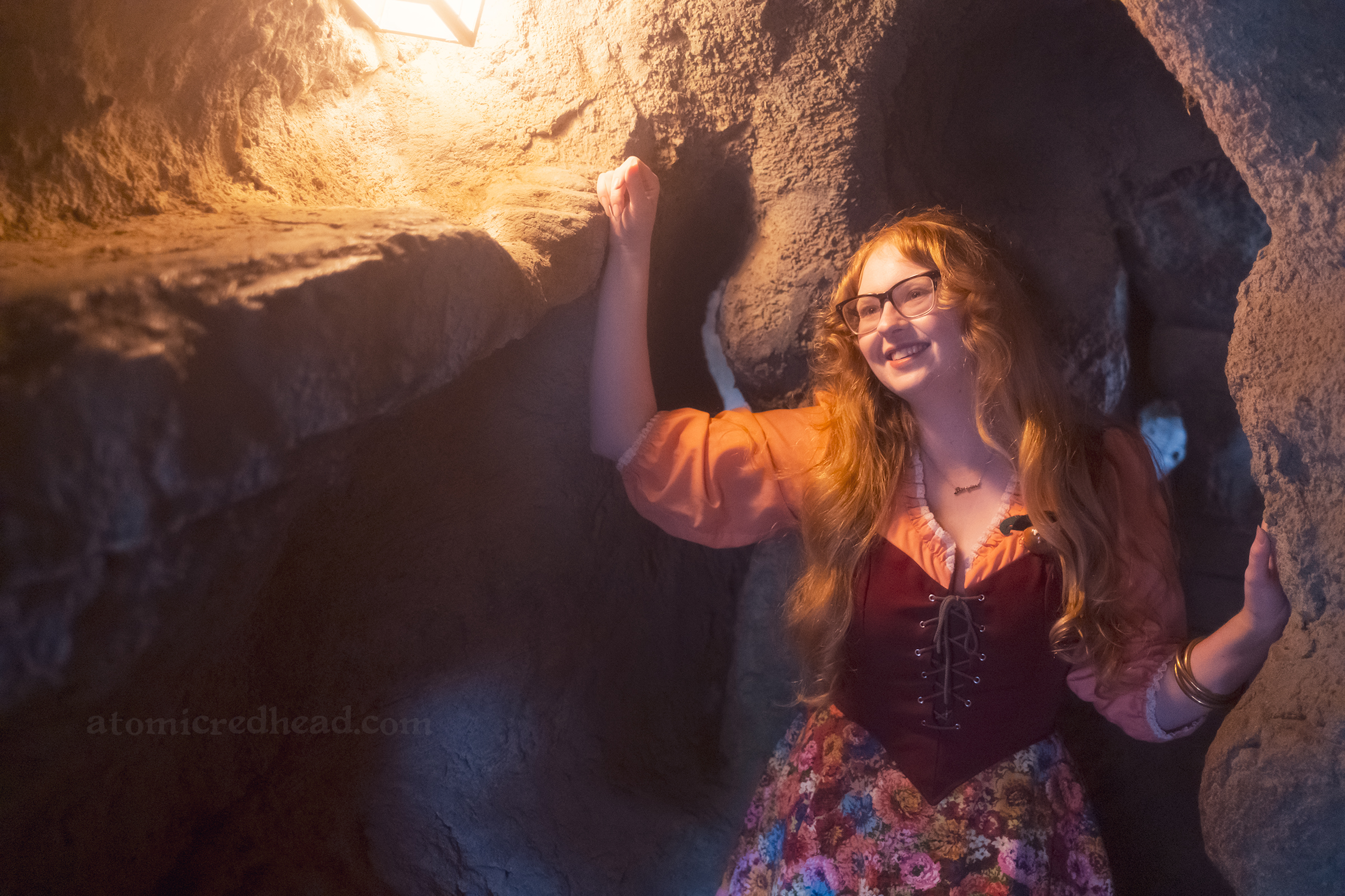 Myself standing in a cave with a light on the left, wearing a copper crown, dress with coral sleeves, maroon lace up bodice, and short floral skirt.