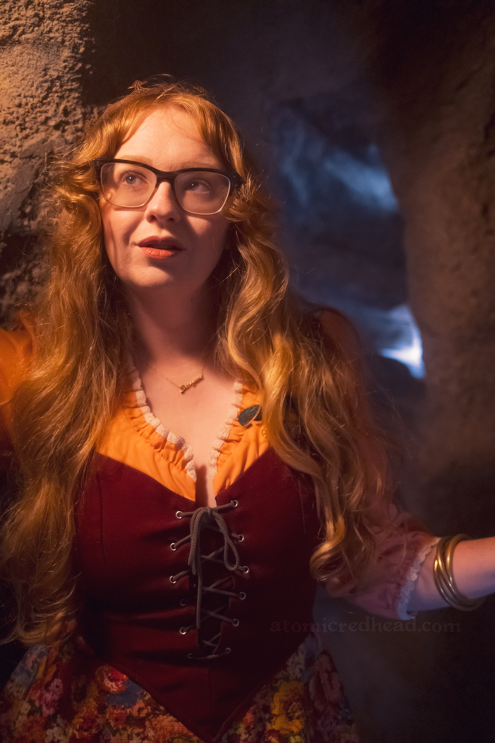 Myself standing in a cave with a light on the left, wearing a copper crown, dress with coral sleeves, maroon lace up bodice, and short floral skirt.