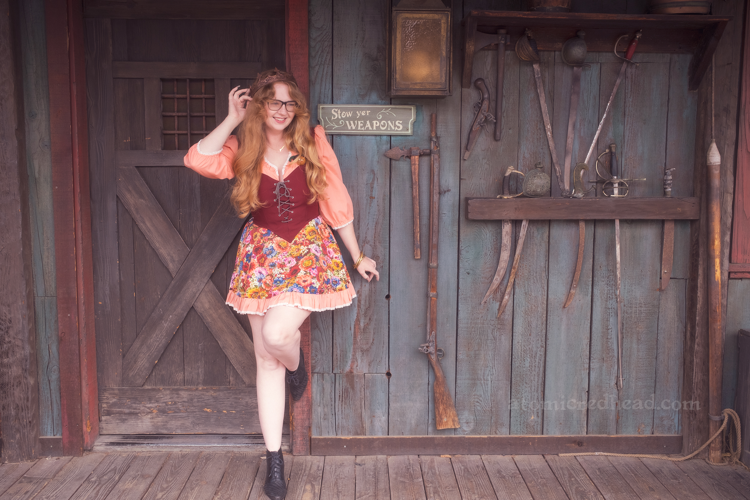 Myself standing against Lafitte Tavern, wearing a copper crown, dress with coral sleeves, maroon lace up bodice, and short floral skirt. On the right a small sign reads "Stowe Yer Weapons" and a rack of swords.
