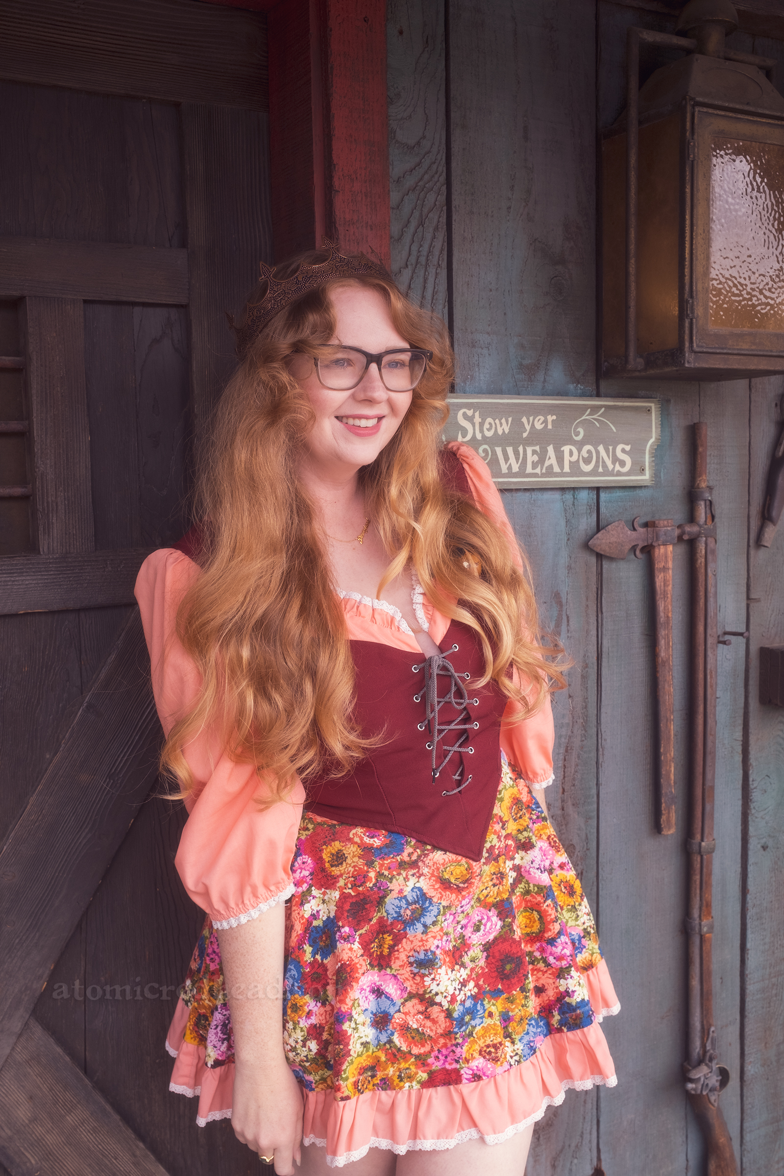 Myself standing against Lafitte Tavern, wearing a copper crown, dress with coral sleeves, maroon lace up bodice, and short floral skirt. On the right a small sign reads "Stowe Yer Weapons" and a rack of swords.