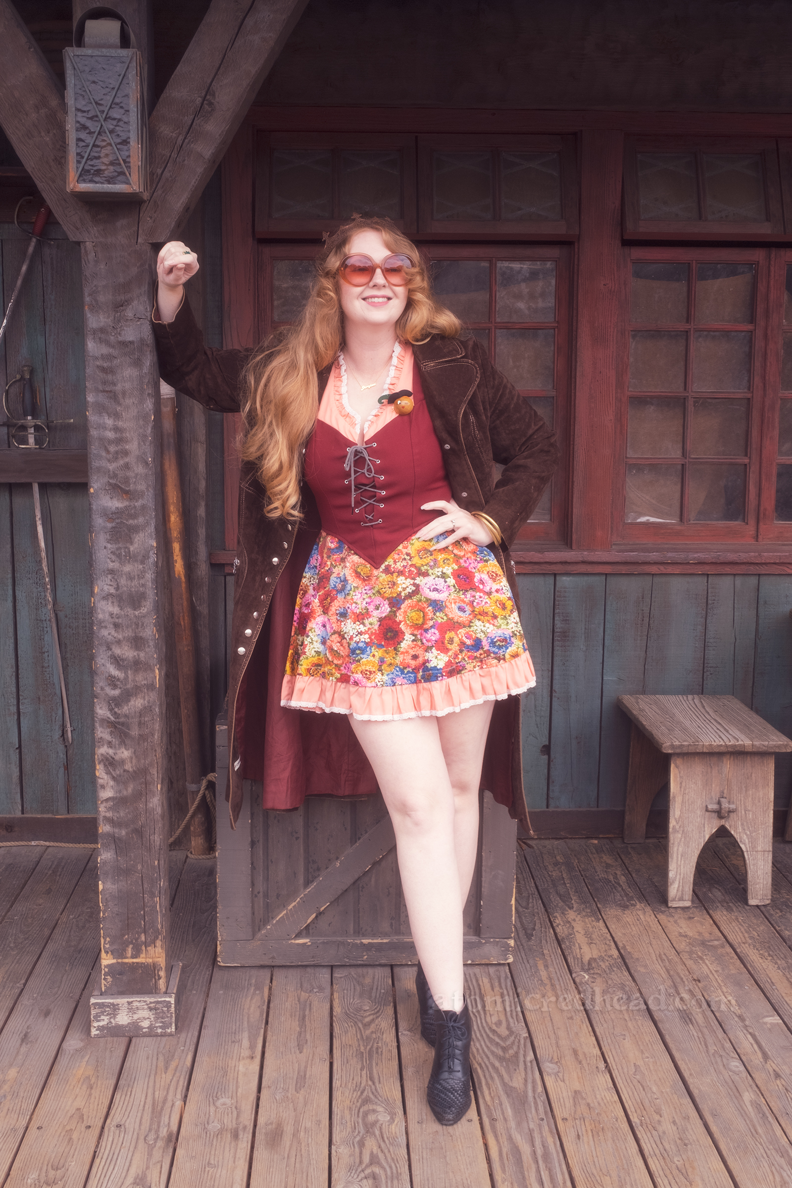 Myself standing against Lafitte Tavern, wearing a copper crown, dress with coral sleeves, maroon lace up bodice, and short floral skirt, with a long brown coat over. On the right a small sign reads "Stowe Yer Weapons" and a rack of swords.