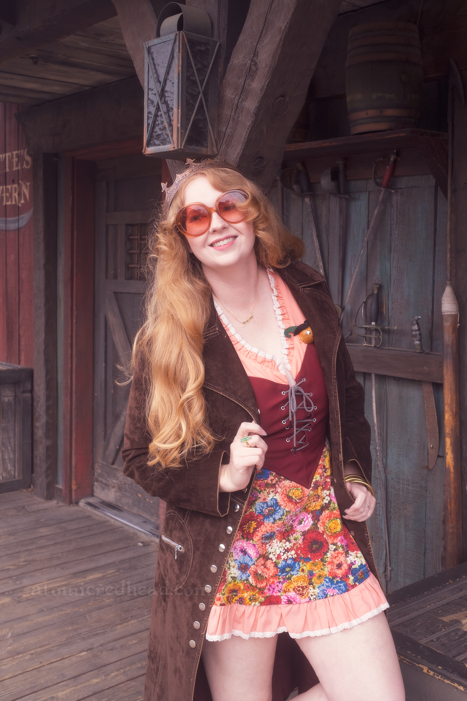 Myself standing against Lafitte Tavern, wearing a copper crown, dress with coral sleeves, maroon lace up bodice, and short floral skirt, with a long brown coat over. On the right a small sign reads "Stowe Yer Weapons" and a rack of swords.