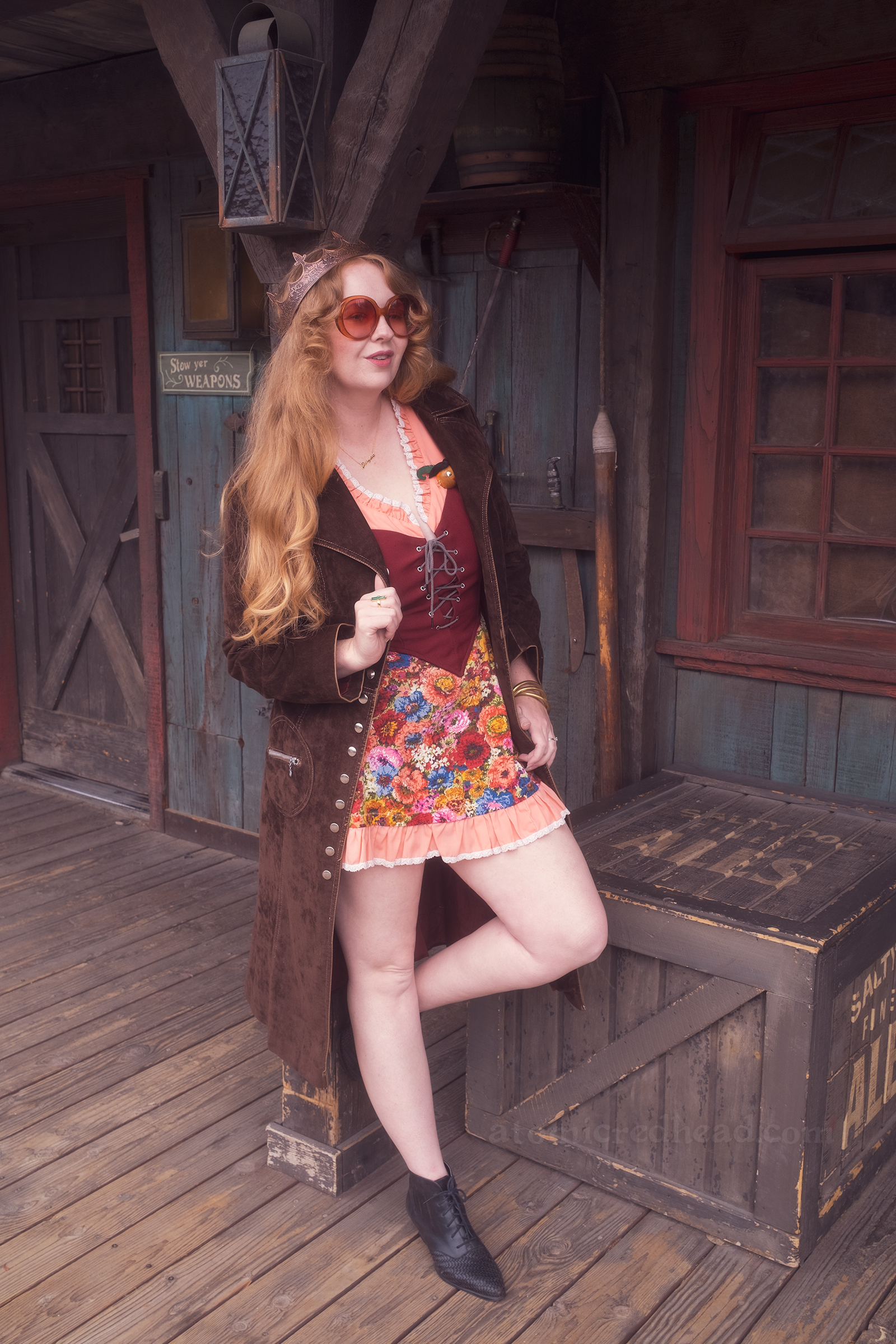 Myself standing against Lafitte Tavern, wearing a copper crown, dress with coral sleeves, maroon lace up bodice, and short floral skirt, with a long brown coat over. On the right a small sign reads "Stowe Yer Weapons" and a rack of swords.