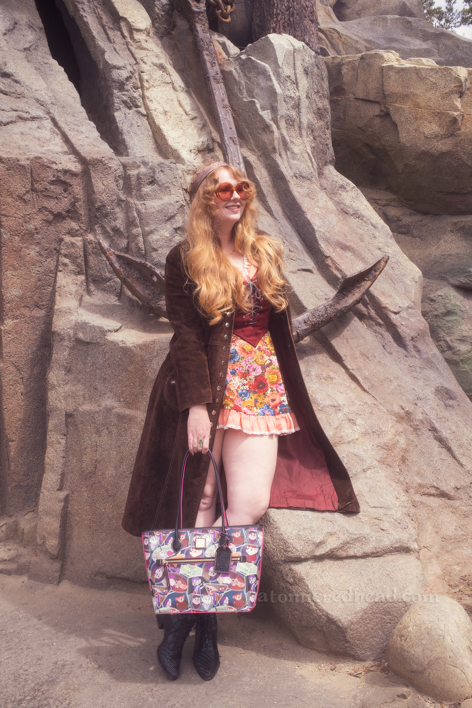 Myself leaning against a rock that has a giant anchor on it, wearing a copper crown, dress with coral sleeves, maroon lace up bodice, and short floral skirt, with a long brown coat over. 