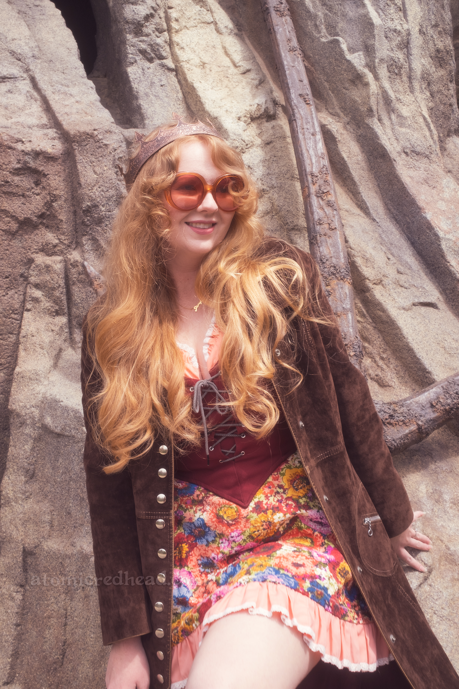 Myself leaning against a rock that has a giant anchor on it, wearing a copper crown, dress with coral sleeves, maroon lace up bodice, and short floral skirt, with a long brown coat over. 