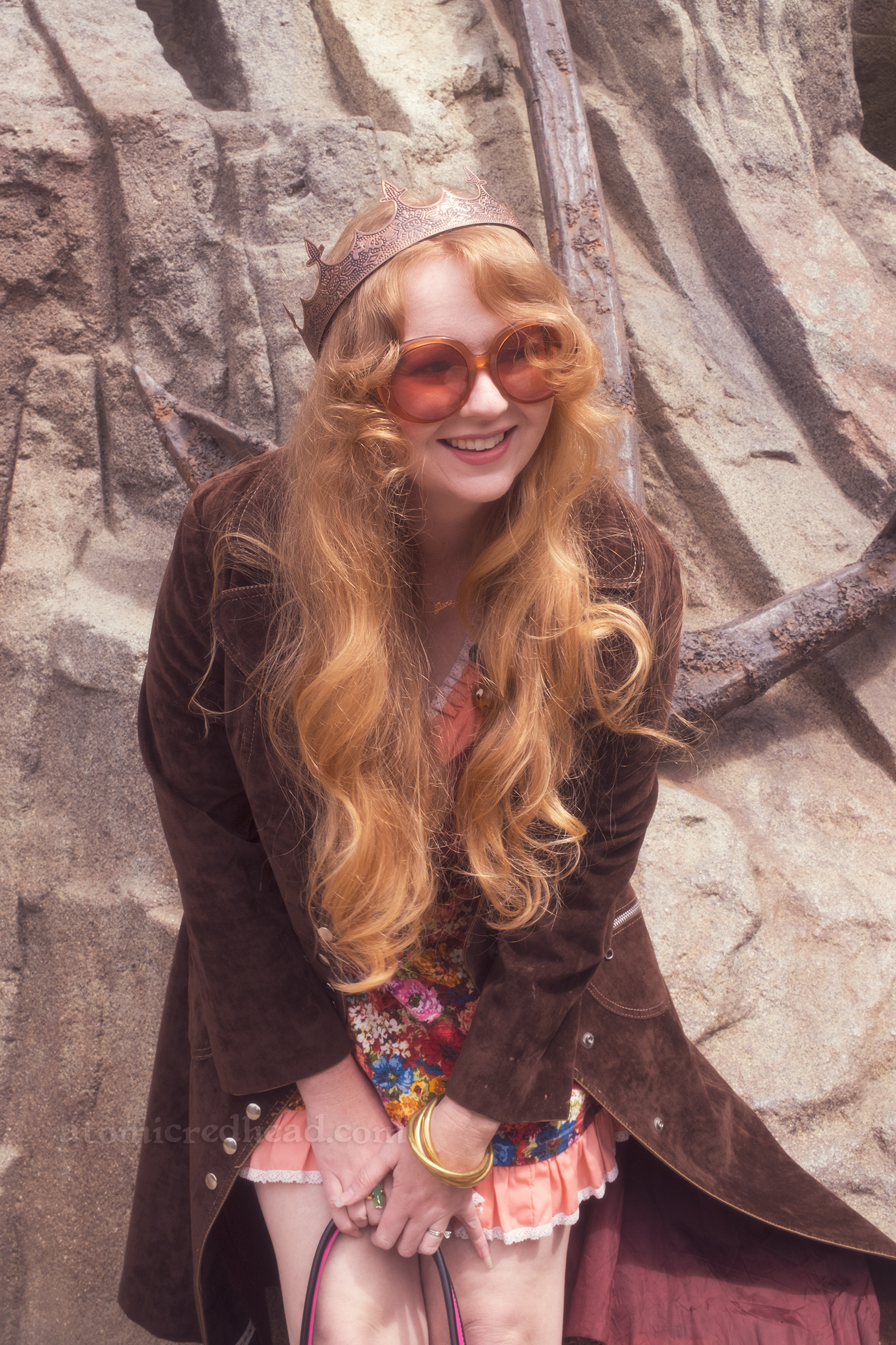 Myself leaning against a rock that has a giant anchor on it, wearing a copper crown, dress with coral sleeves, maroon lace up bodice, and short floral skirt, with a long brown coat over. 