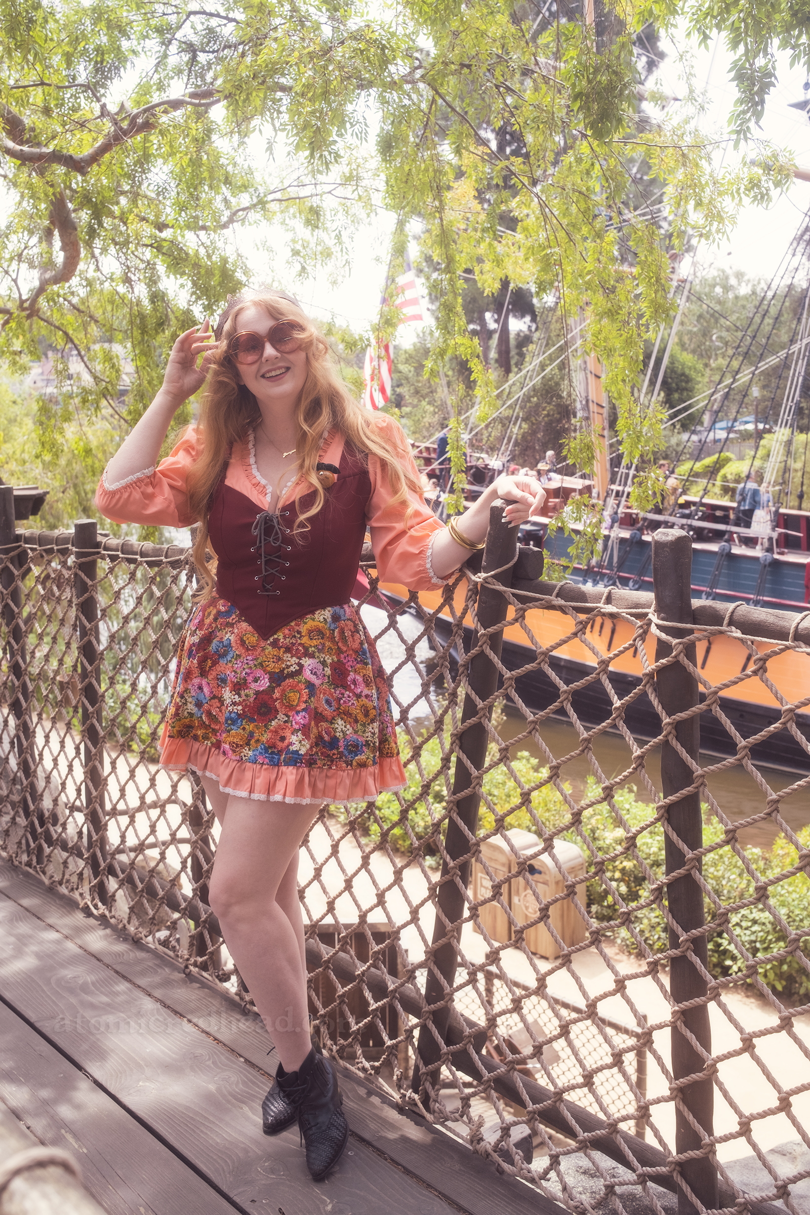 Myself standing, leaning against a rope railing, wearing a copper crown, dress with coral sleeves, maroon lace up bodice, and short floral skirt.