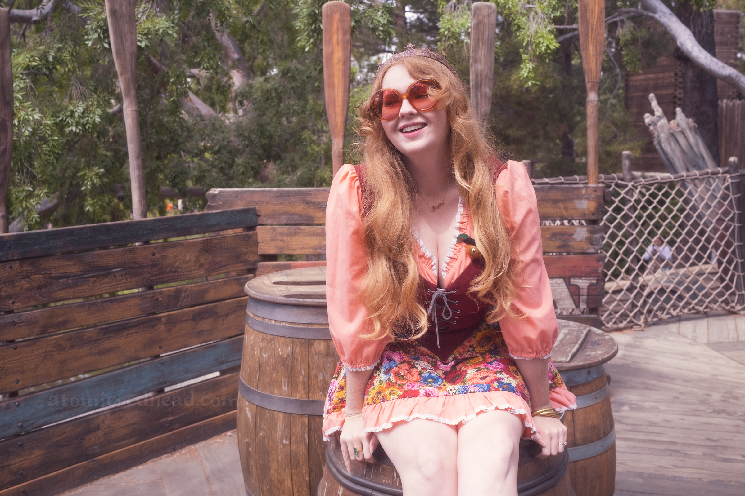 Myself, sitting on a barrel, wearing a copper crown, dress with coral sleeves, maroon lace up bodice, and short floral skirt.