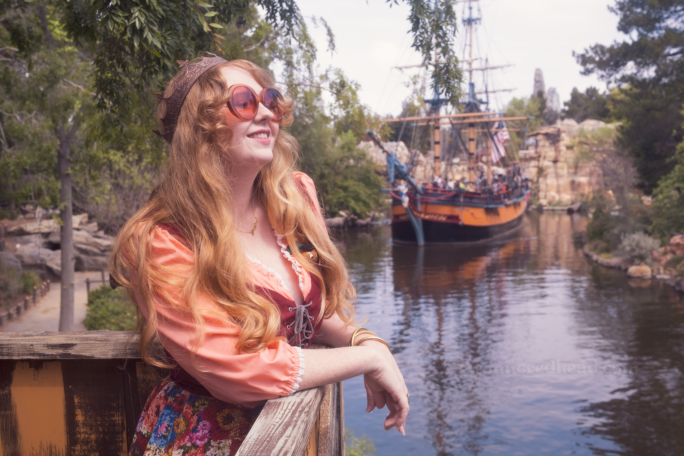 Myself, leaning over a railing, wearing a copper crown, dress with coral sleeves, maroon lace up bodice, and short floral skirt, overlooking the Rivers of America as the sailing ship Columbia comes down the river.